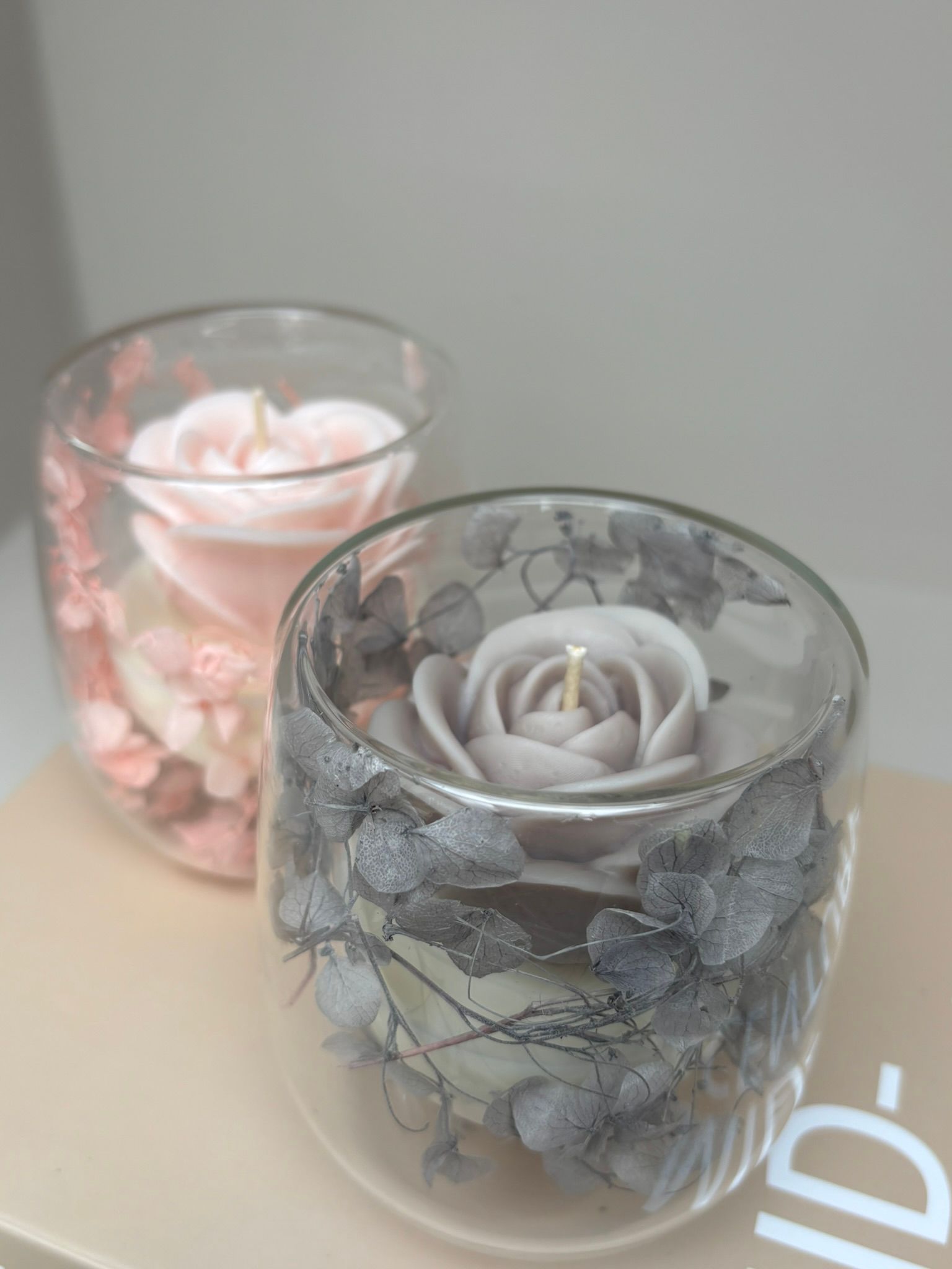 Twee kaarsen van mat glas met bloemmotieven op een beige boek of doos.