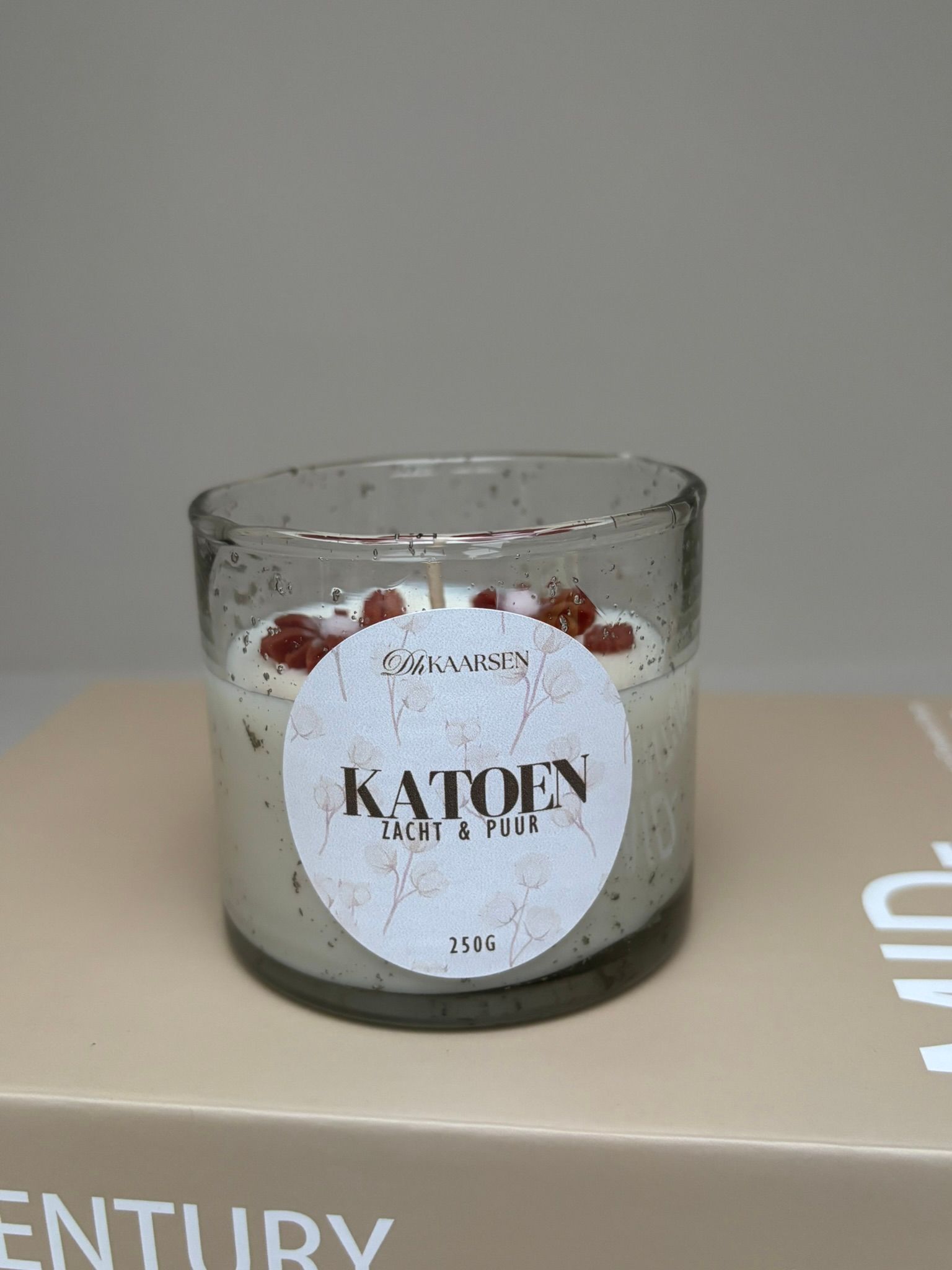 Klein glazen kaarsje met een wit etiket op een beige doosje tegen een grijze achtergrond.