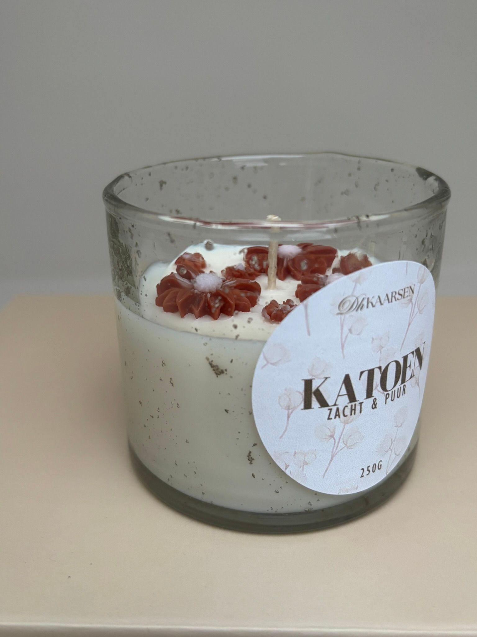 Klein glazen kaarsje met een wit etiket op een beige doosje tegen een grijze achtergrond.