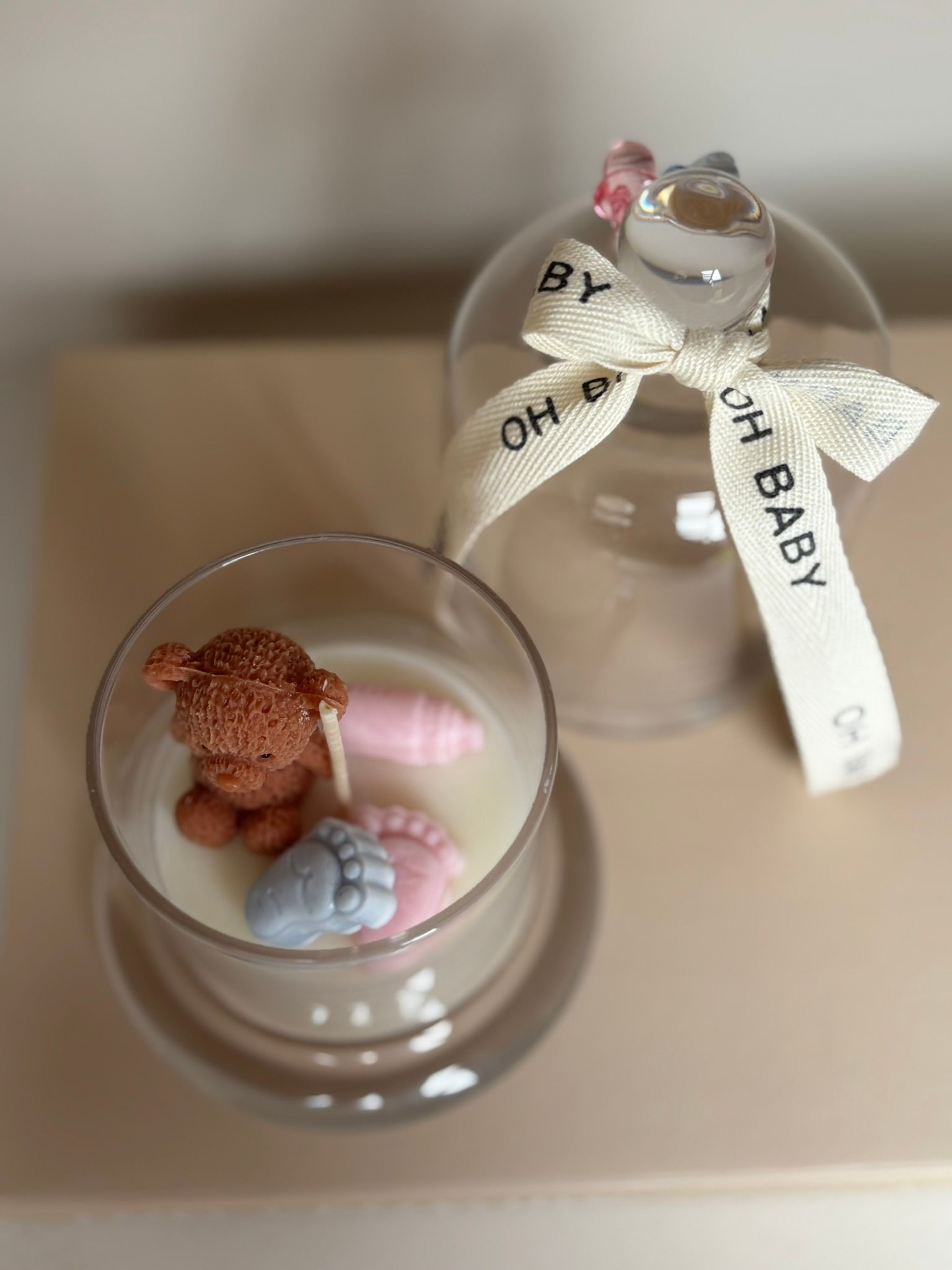 Klein glazen ornament met een teddybeer en pastelkleurige versieringen op een beige ondergrond, vastgebonden met een wit lint. baby