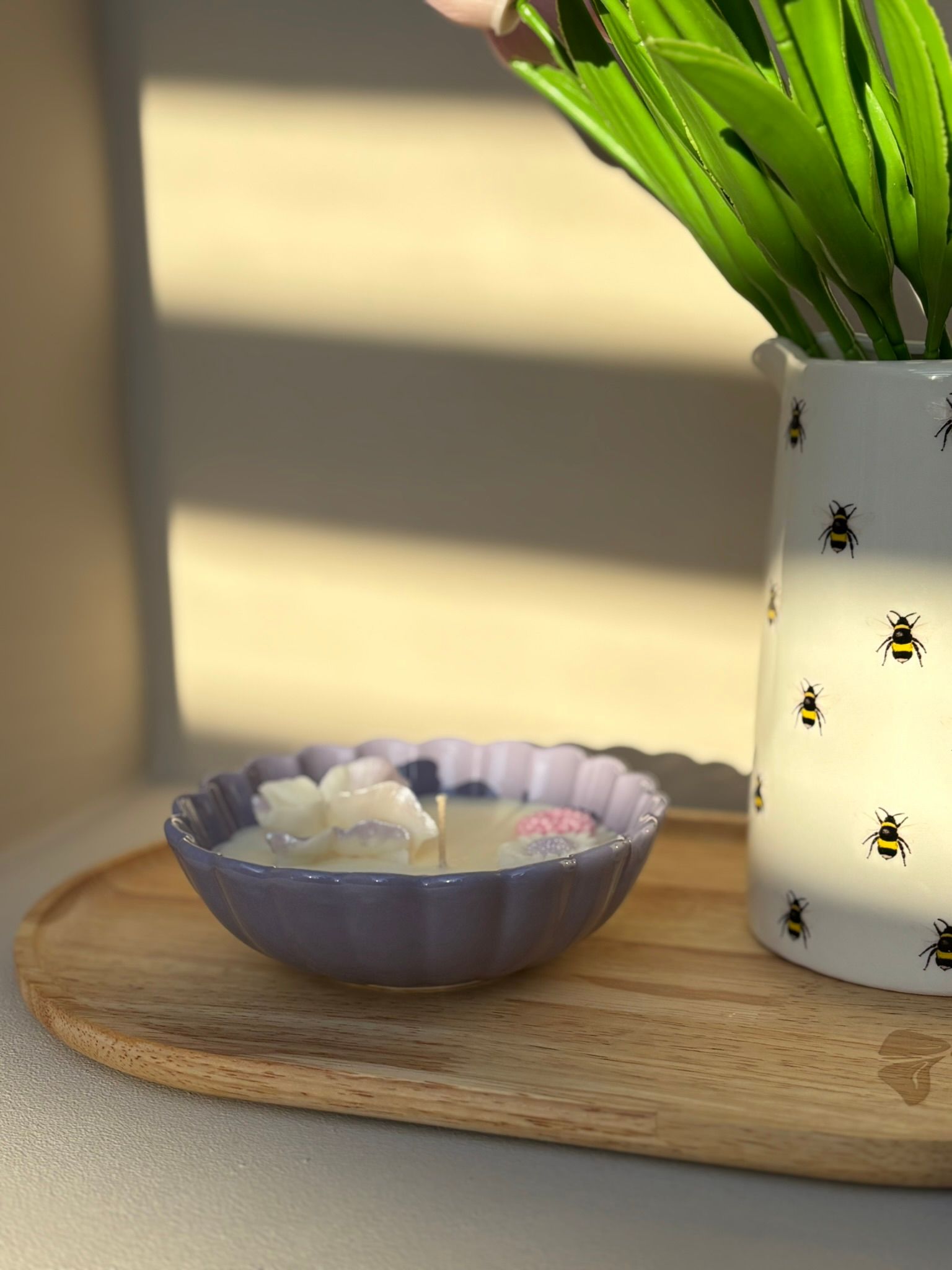 Een witte vaas met groene bladeren staat naast een klein paars schaaltje op een houten dienblad in het warme zonlicht.