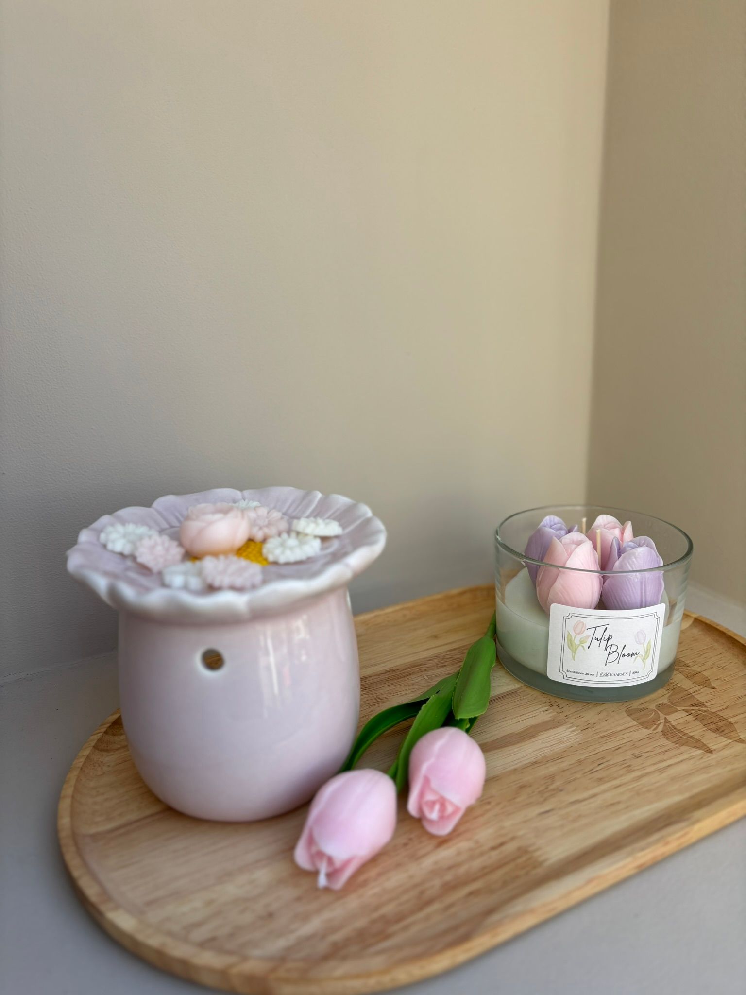 Glazen kaarsenhouder met pastelroze en paarse bloemen op een houten dienblad naast een roze bloem.