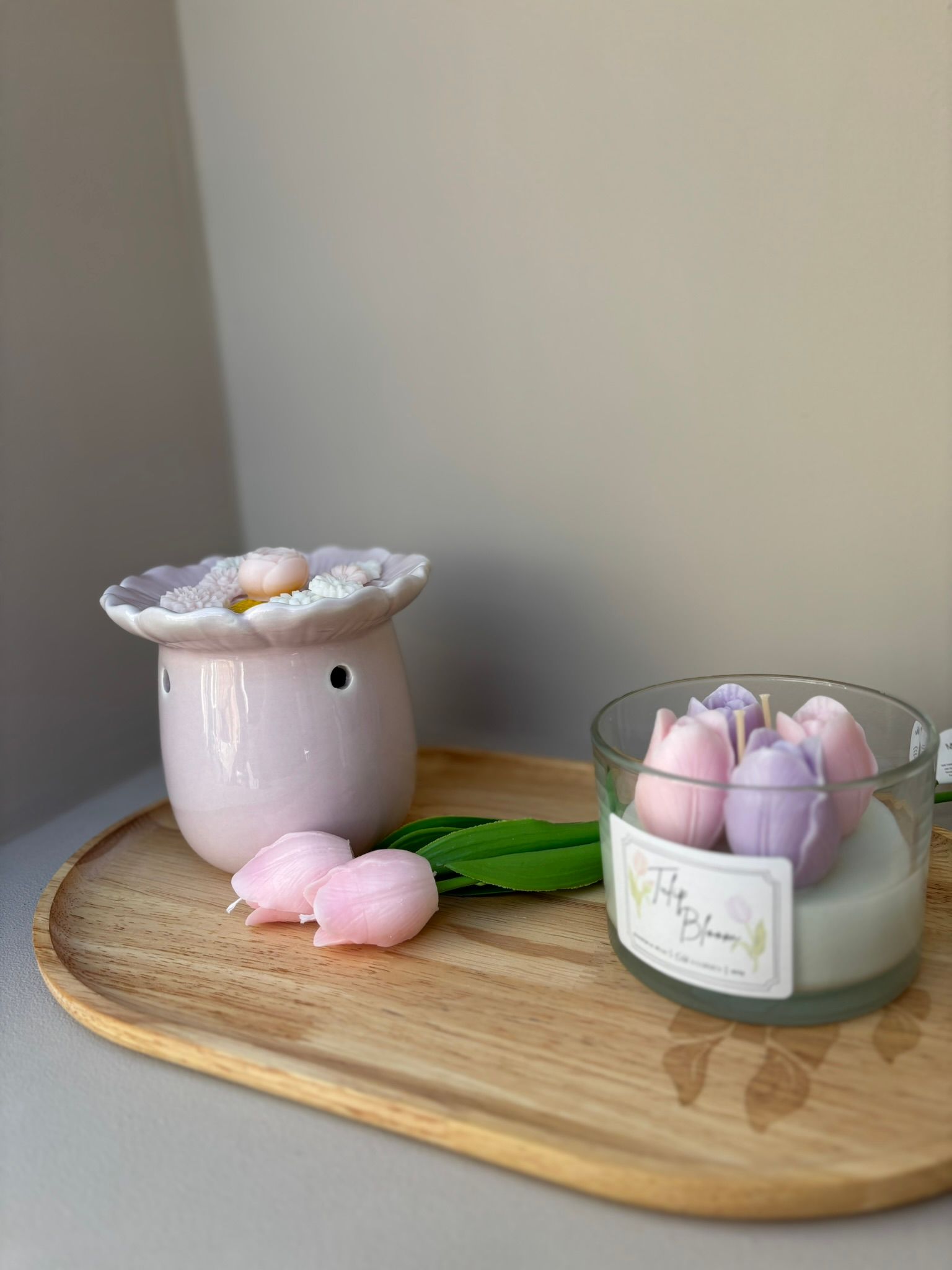 Witte, bloemvormige kaarsenhouder op een houten dienblad met roze tulpen tegen een beige muur.