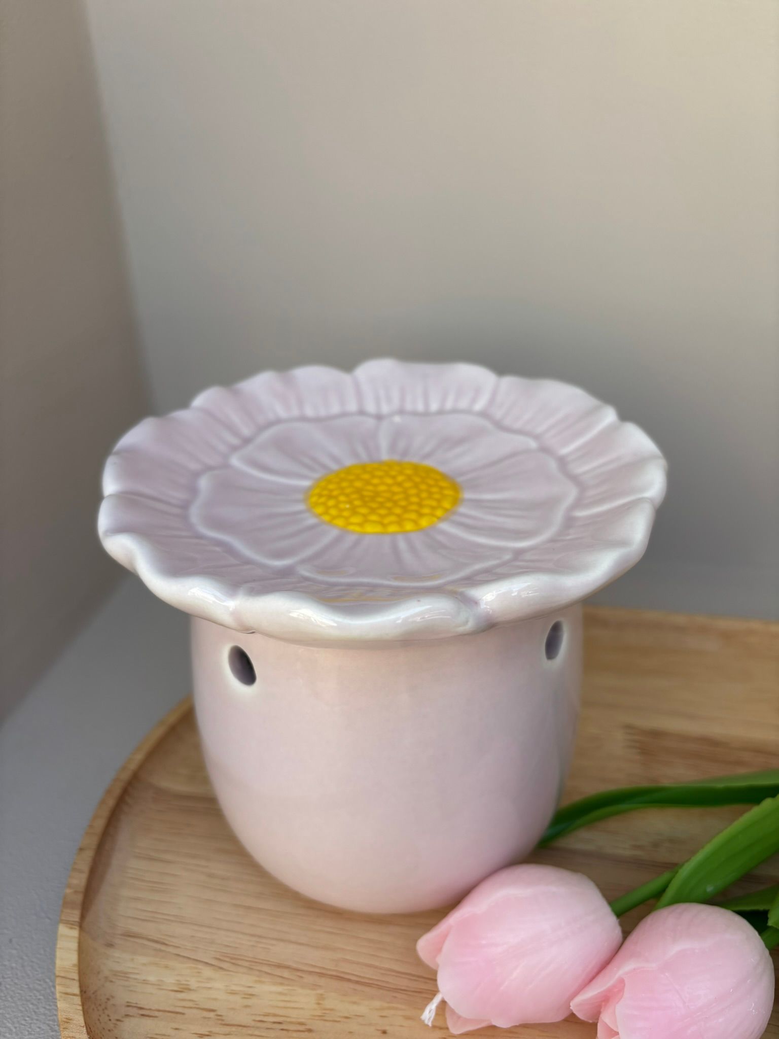 Witte, bloemvormige kaarsenhouder op een houten dienblad met roze tulpen tegen een beige muur.