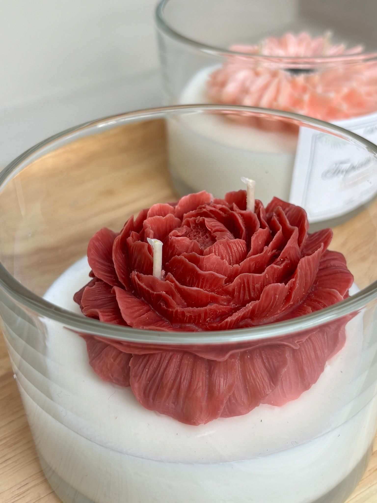 Witte kaars in een glazen pot met een rode bloem op een houten dienblad, met daarachter een tweede, wazige kaars.
