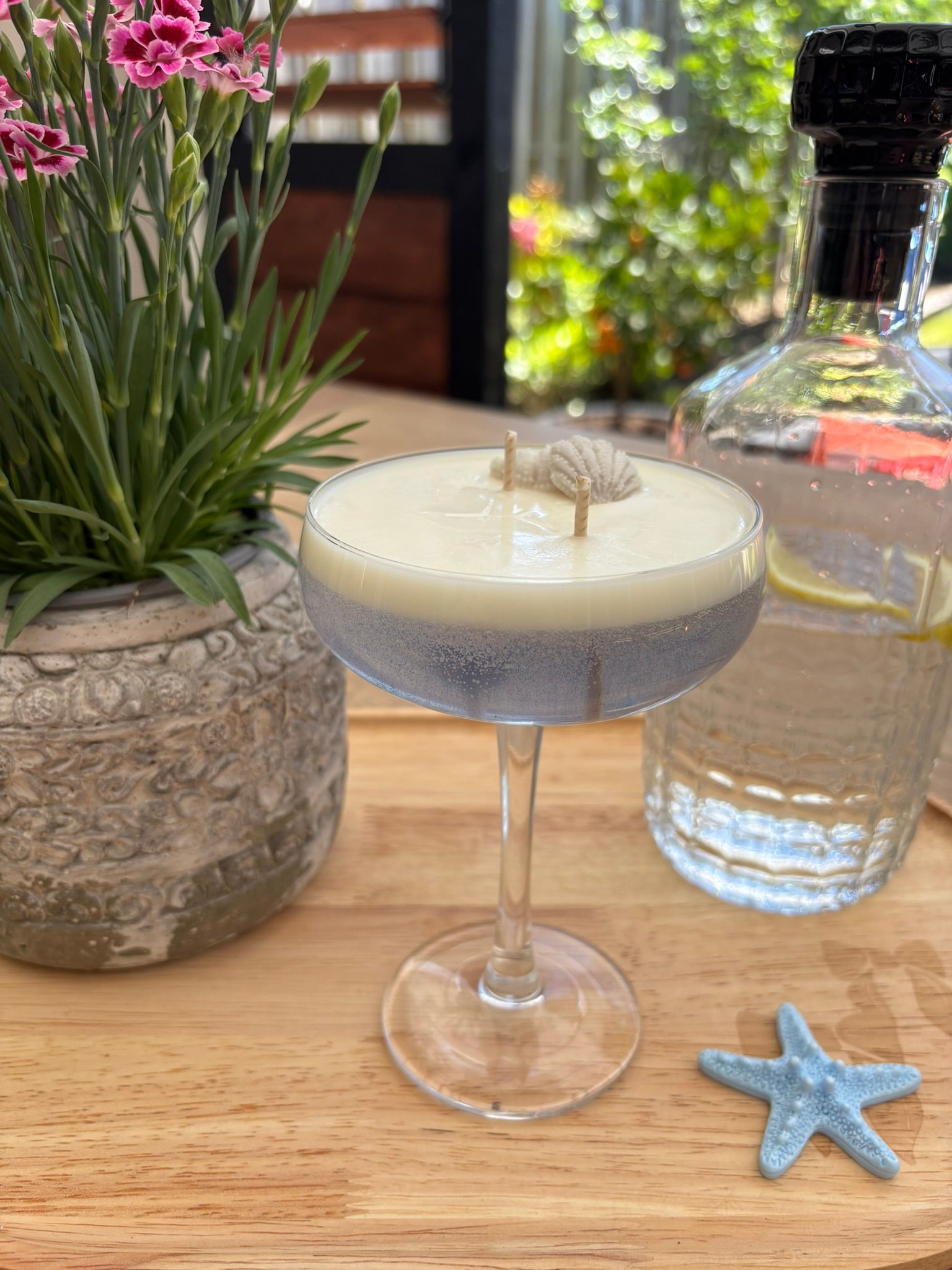 Een cocktailkaars in een coupeglas op een houten tafeltje naast een potplant, een waterfles en een blauwe zeester.