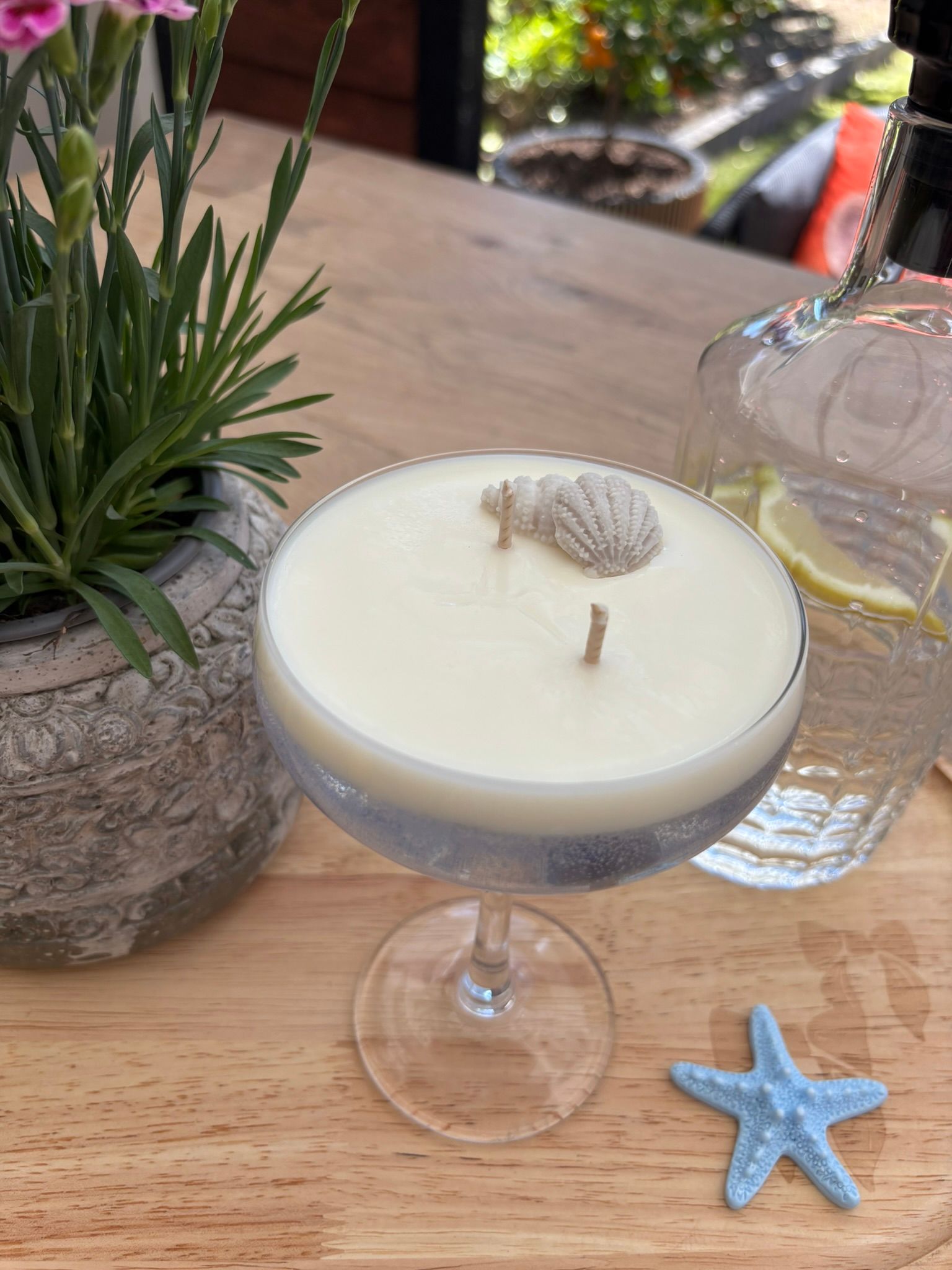 Een cocktailkaars in een coupeglas op een houten tafeltje naast een potplant, een waterfles en een blauwe zeester.