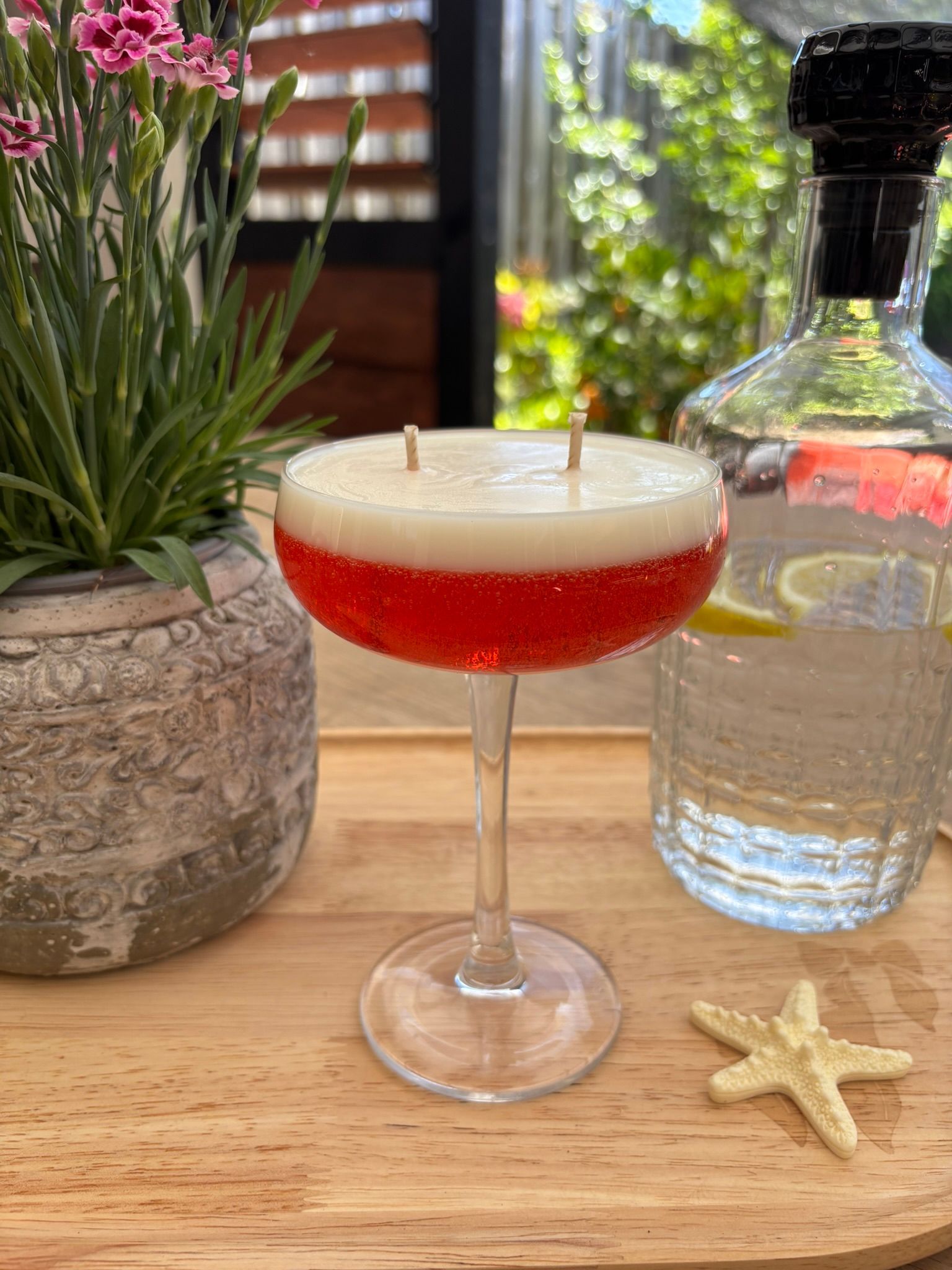 Een rode cocktail in een coupeglas op een houten tafeltje naast een potplant en een fles water, met een zeester erop.