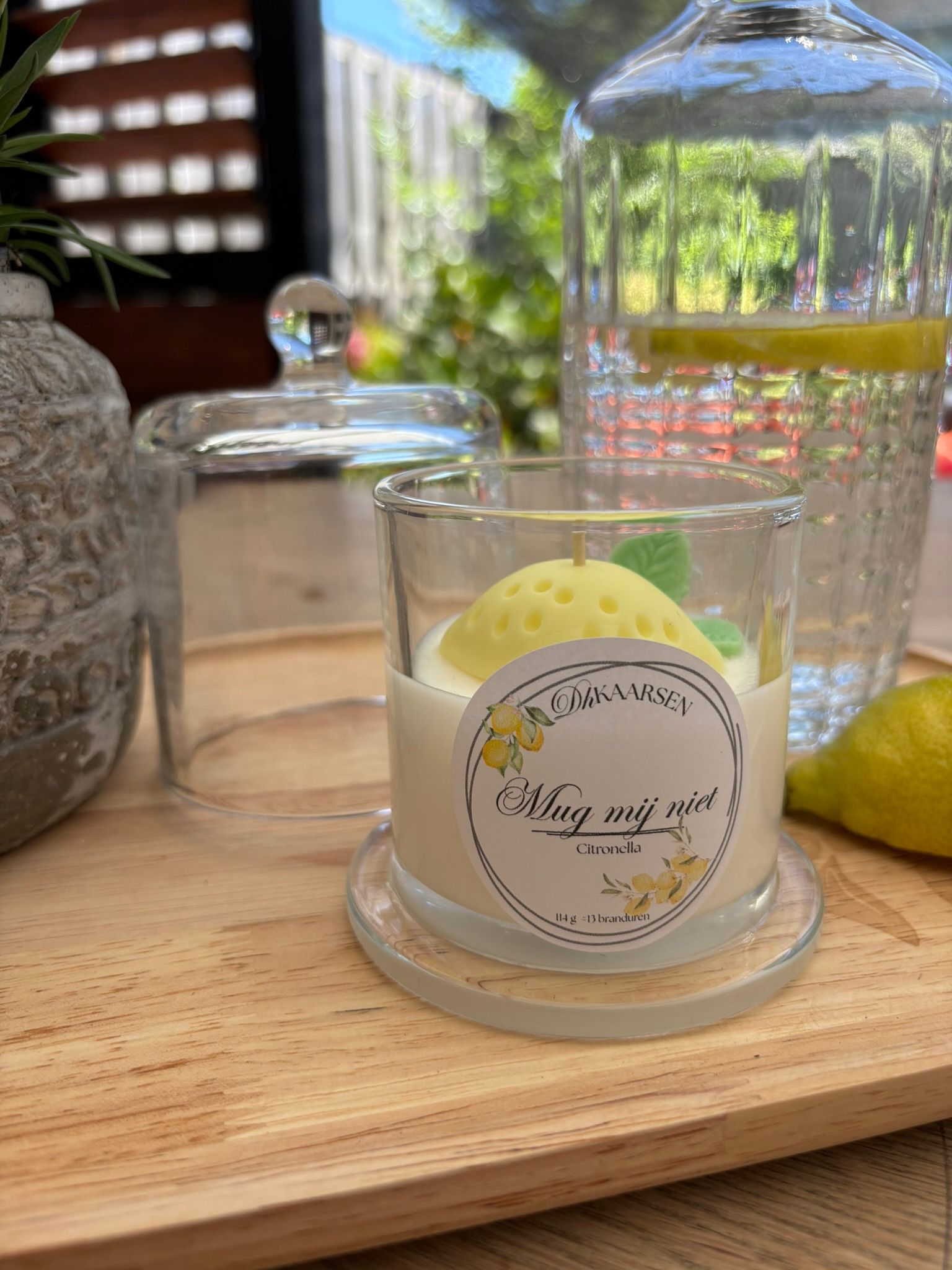 Een klein kaarsenpotje met gele was staat op een houten tafeltje naast glazen stolpen en een waterkan bij het raam.