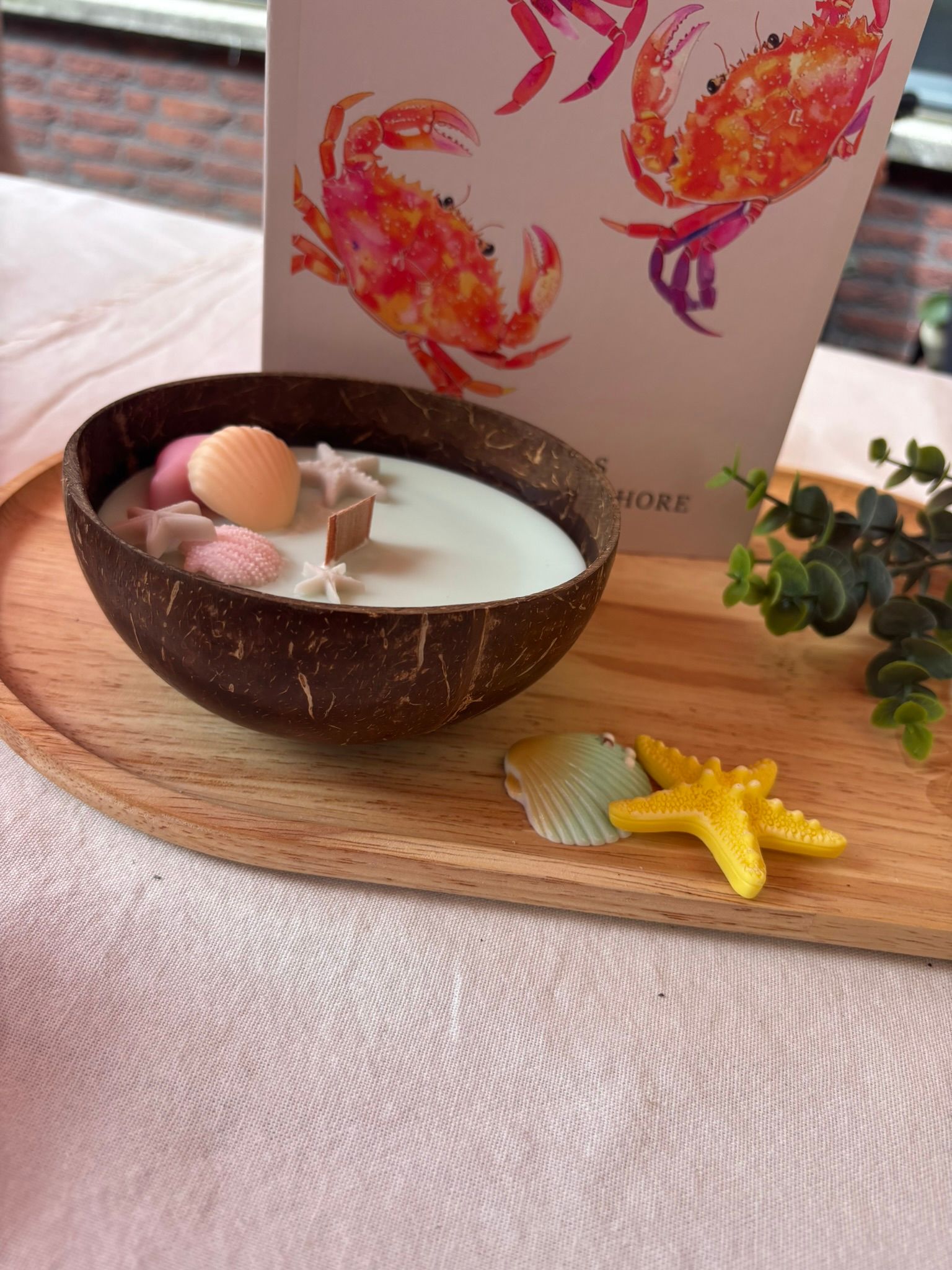 Een kaars in een kokosnootschaal met schelp- en zeesterrenversiering op een roze tafelkleed, naast een kaartje met bloemen.