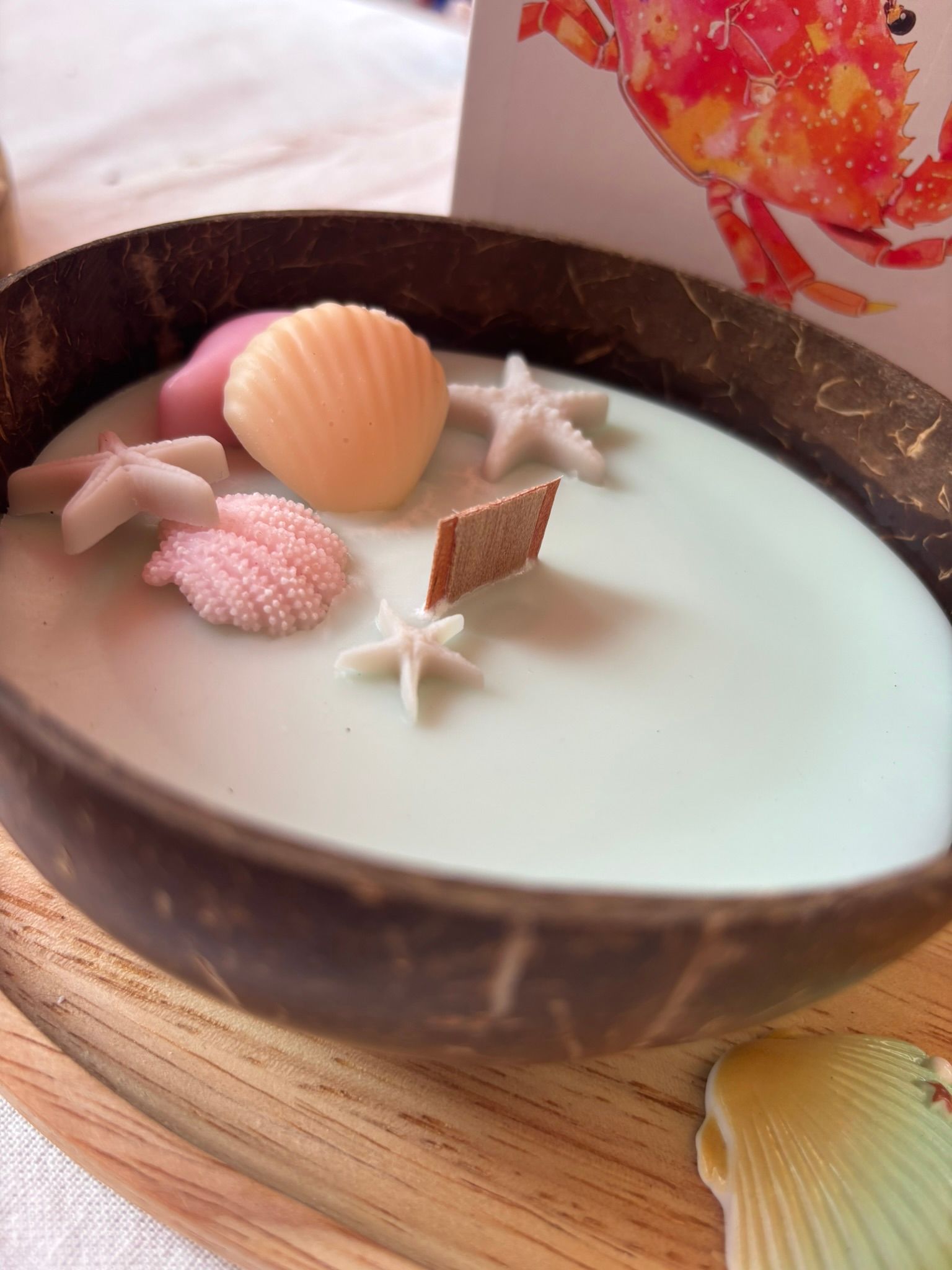 Een kaars in een kokosnootschaal met schelp- en zeesterrenversiering op een roze tafelkleed, naast een kaartje met bloemen.