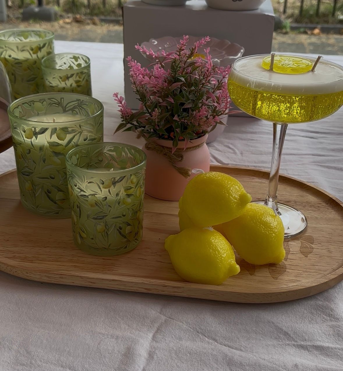 Groene glazen met patroon, een citroengarnituur, roze bloemen en een gele cocktail op een dienblad in de buitenlucht.