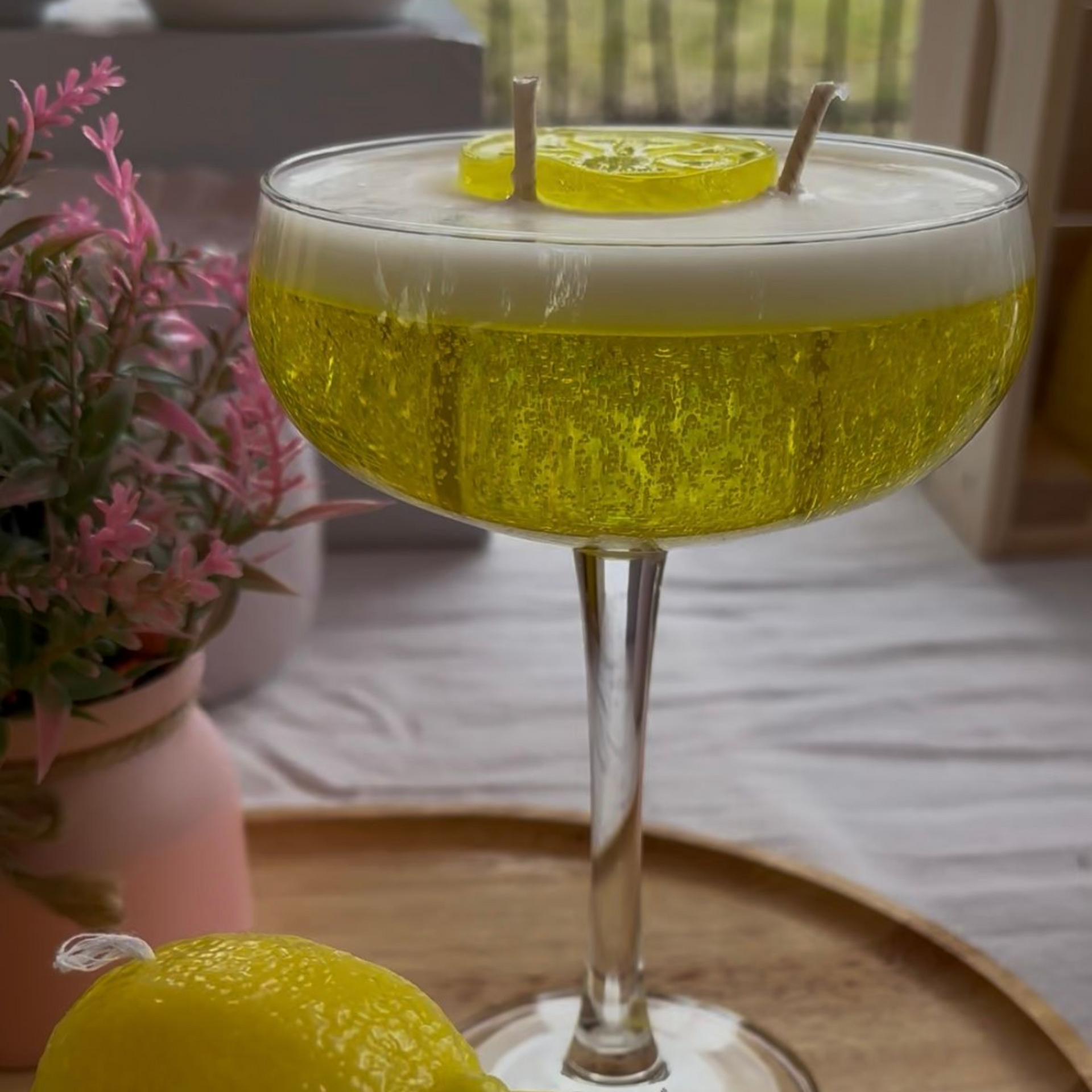 Groene glazen met patroon, een citroengarnituur, roze bloemen en een gele cocktail op een dienblad in de buitenlucht.