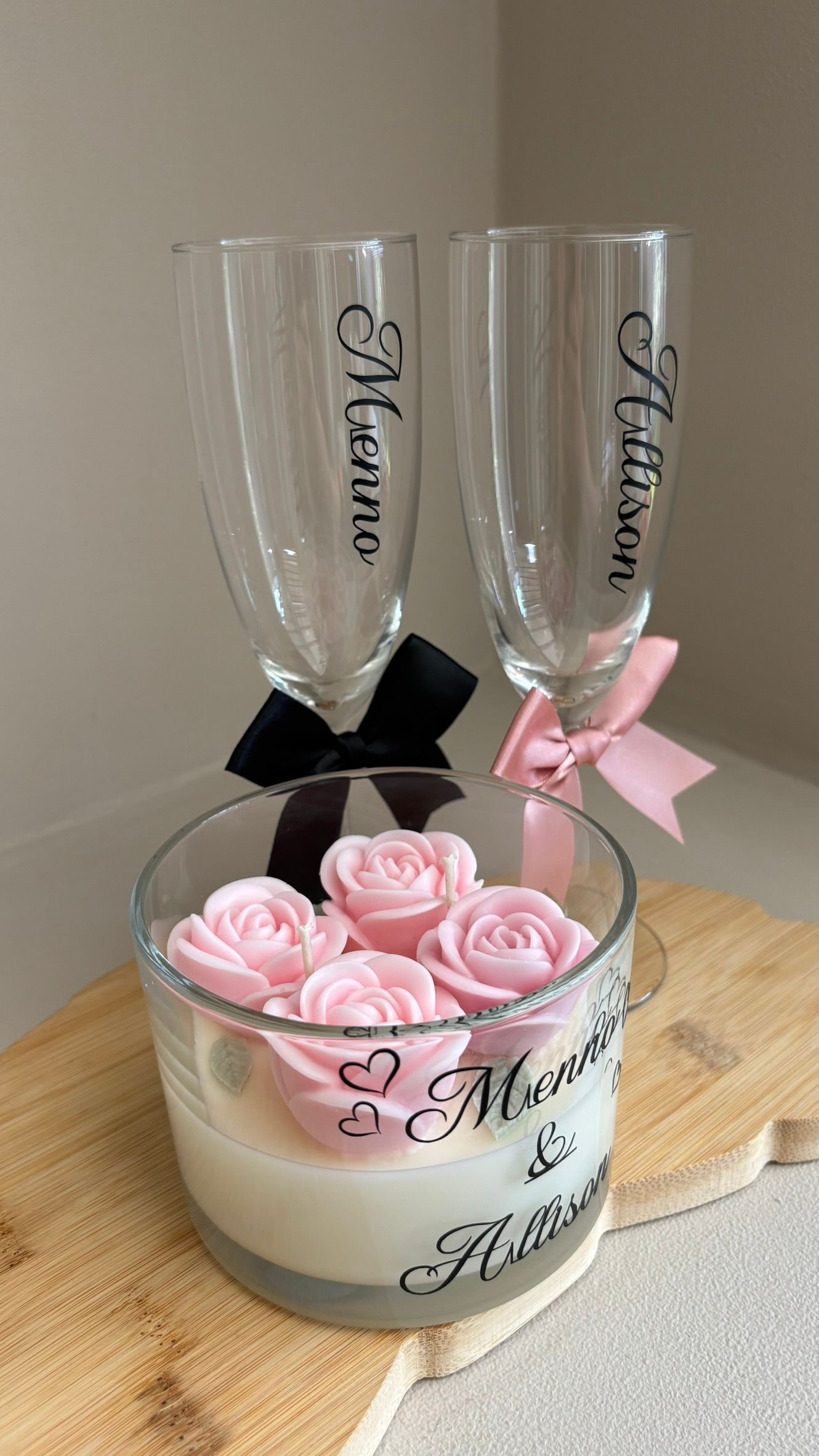 Roze rozen in een versierde kaarsenhouder met twee gepersonaliseerde champagneglazen, vastgebonden met zwarte en roze linten.
