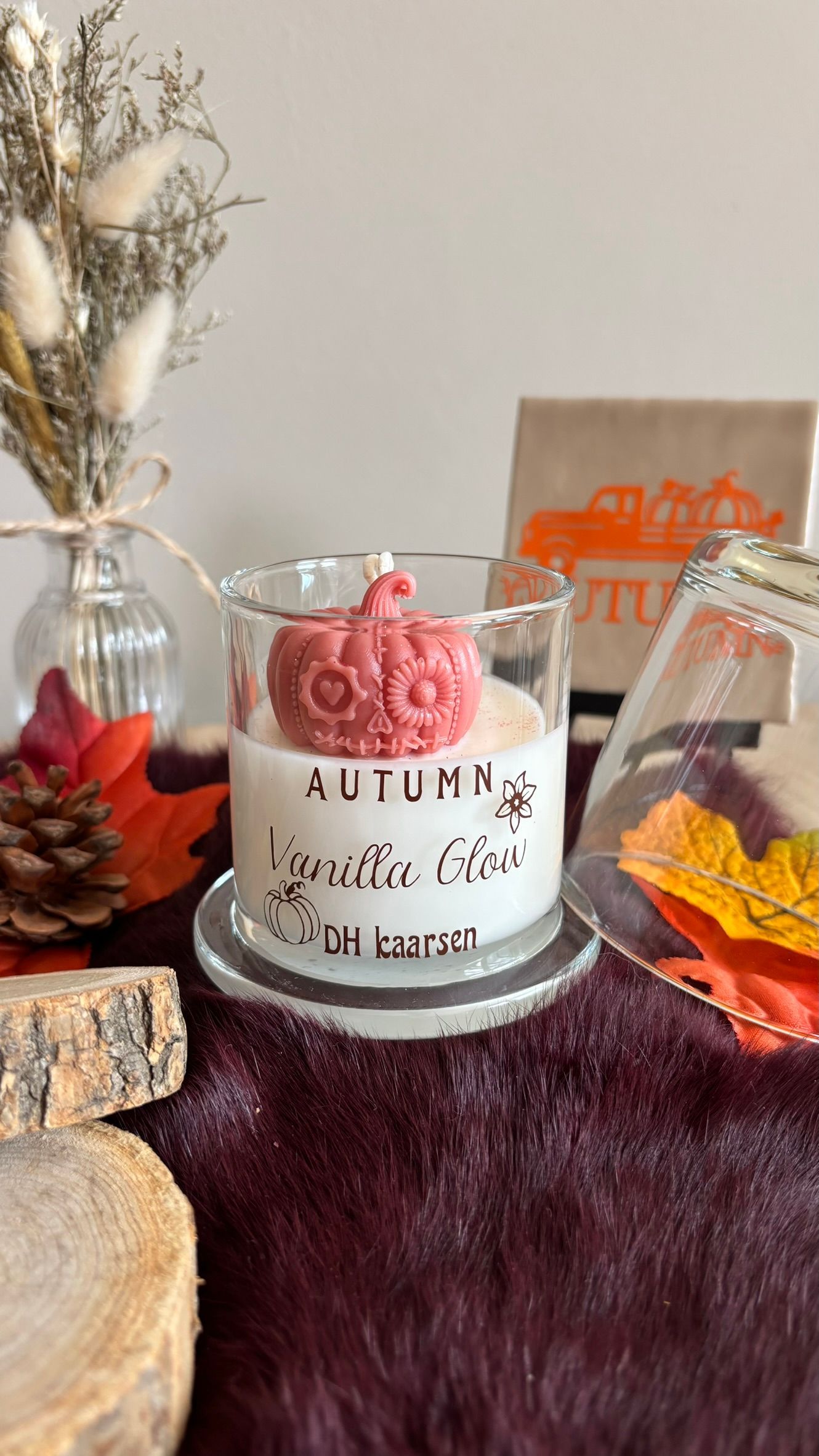 Een kaars met herfstthema in een glazen pot met roze wassmelt op een bordeauxrode, zachte ondergrond, naast dennenappels en bladeren.