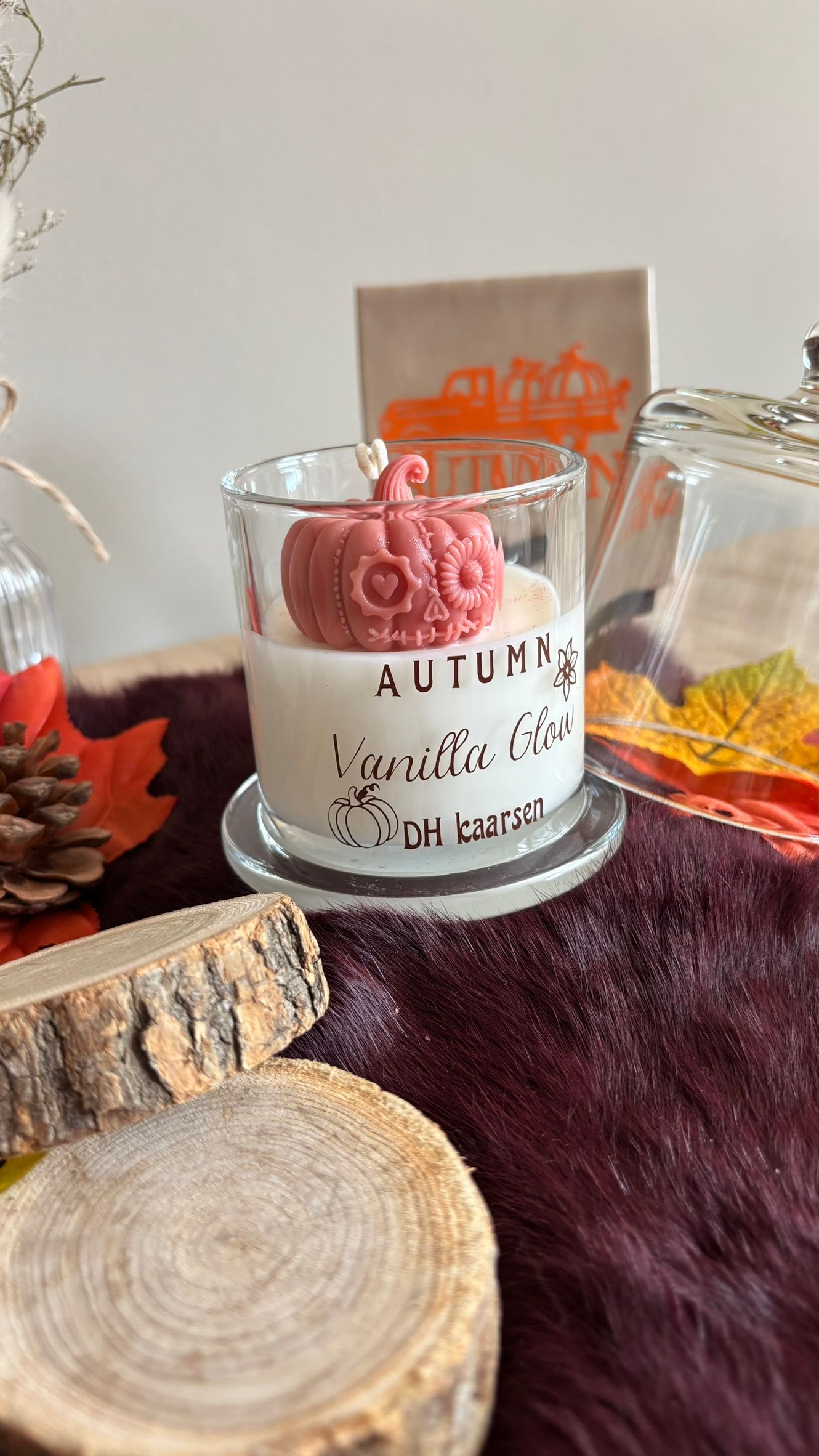 Een kaars met herfstthema in een glazen pot met roze wassmelt op een bordeauxrode, zachte ondergrond, naast dennenappels en bladeren.