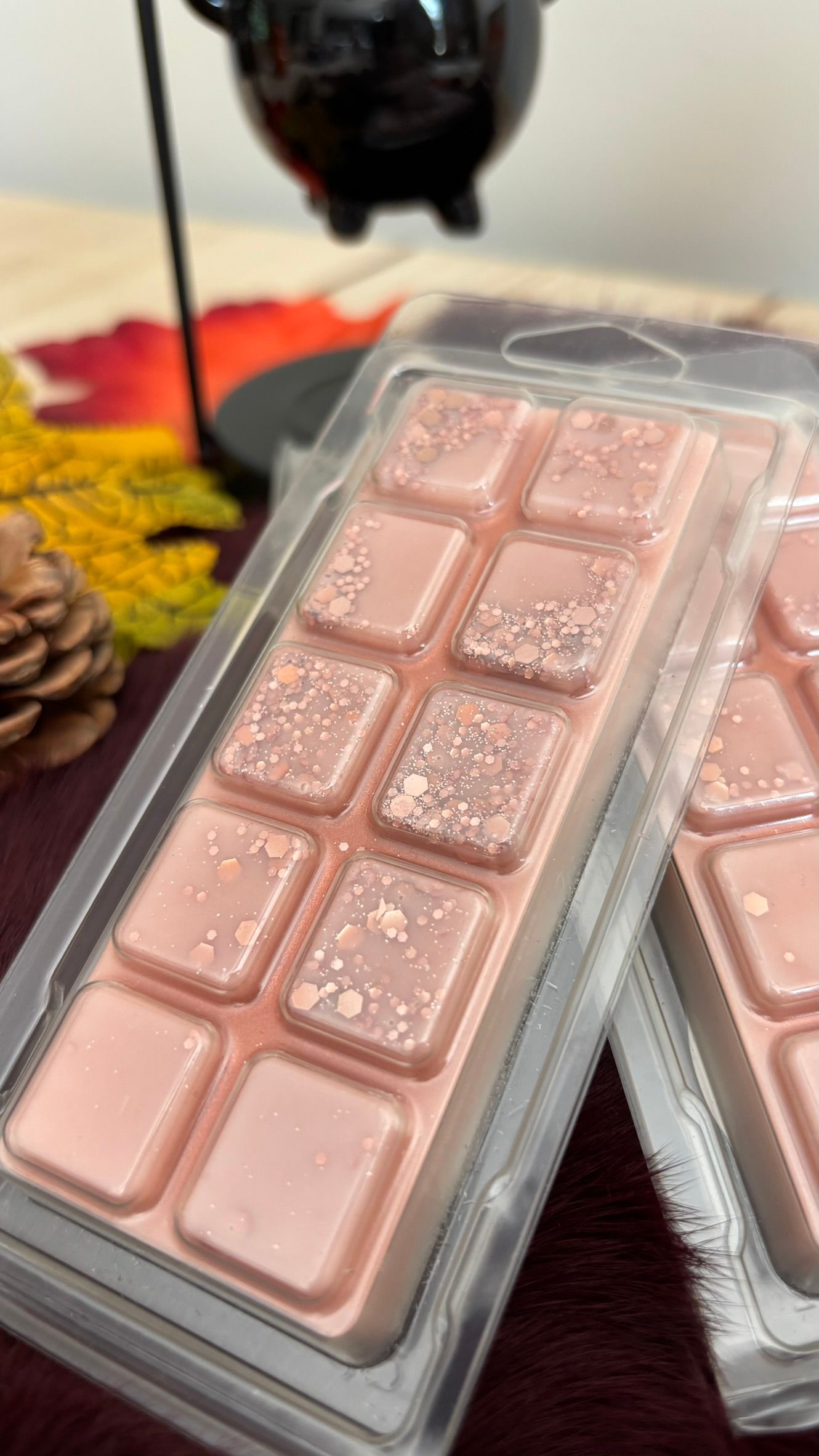Drie roze chocoladerepen in grijze verpakkingen op een bordeauxrode deken naast een zwarte theepot en een dennenappel.