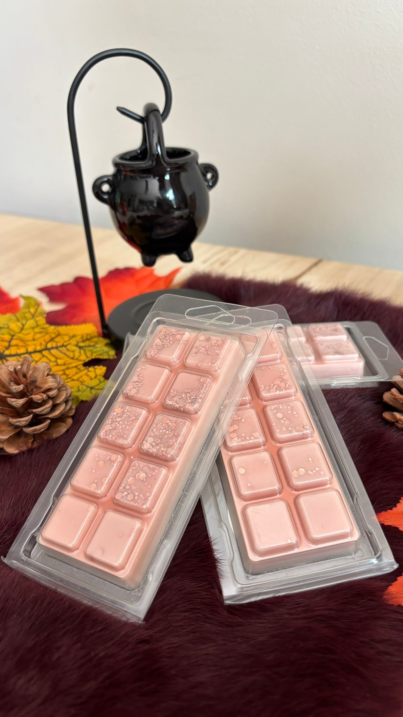 Drie roze chocoladerepen in grijze verpakkingen op een bordeauxrode deken naast een zwarte theepot en een dennenappel.