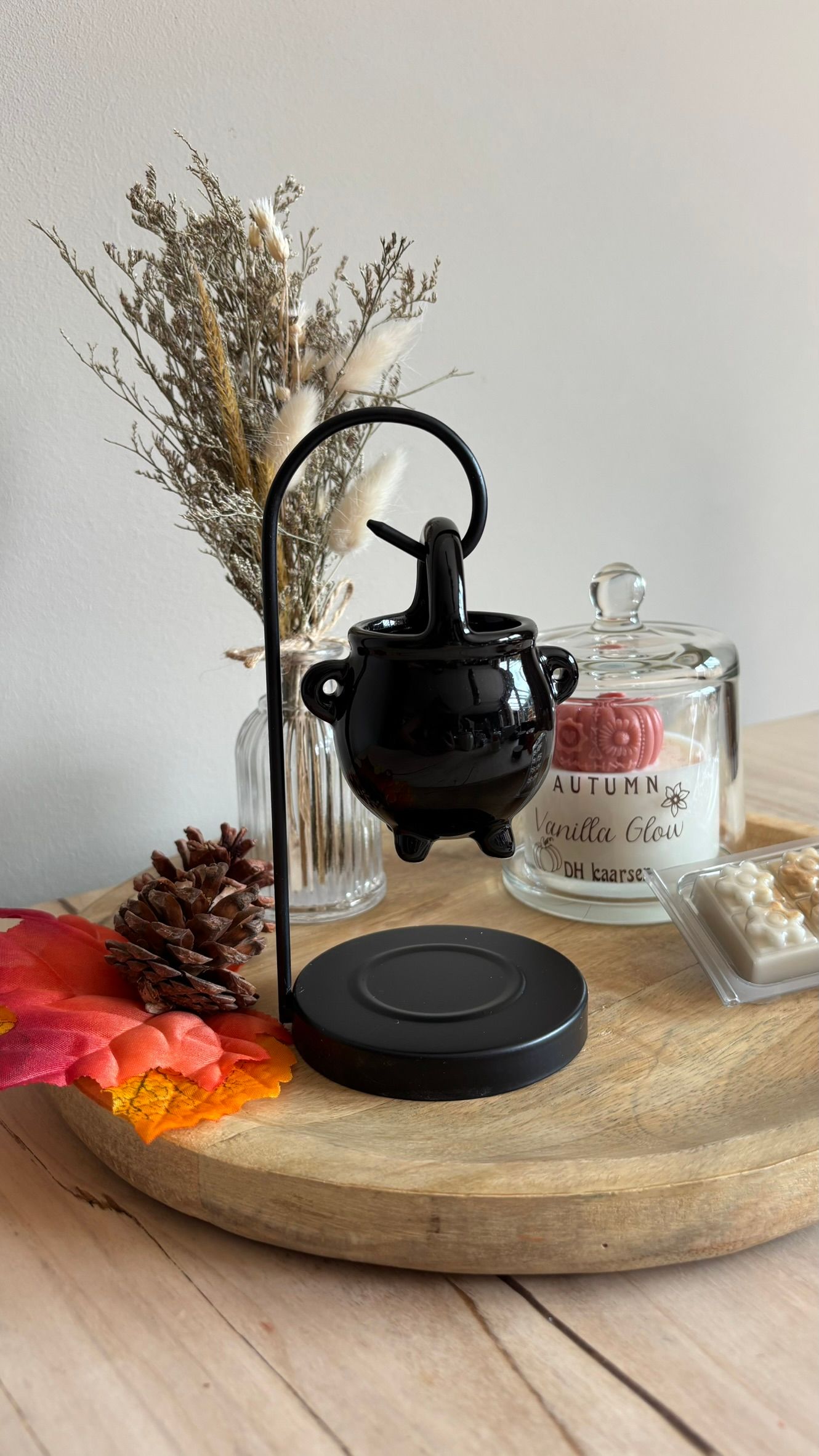Een zwart miniatuurtheepotje op een houten dienblad met gedroogde bloemen, een glazen potje en een rode herfstbladdecoratie.