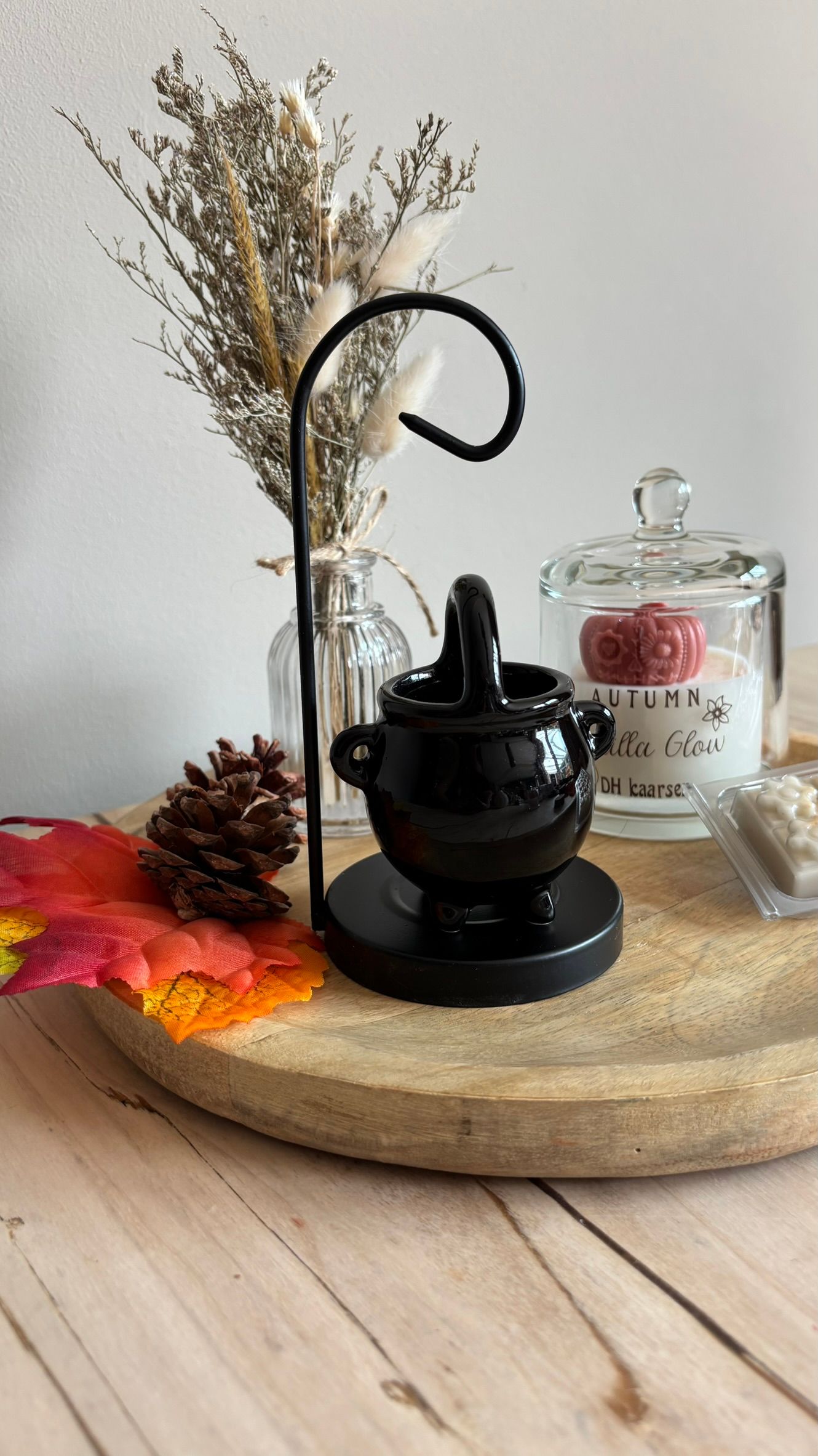 Een zwart miniatuurtheepotje op een houten dienblad met gedroogde bloemen, een glazen potje en een rode herfstbladdecoratie.