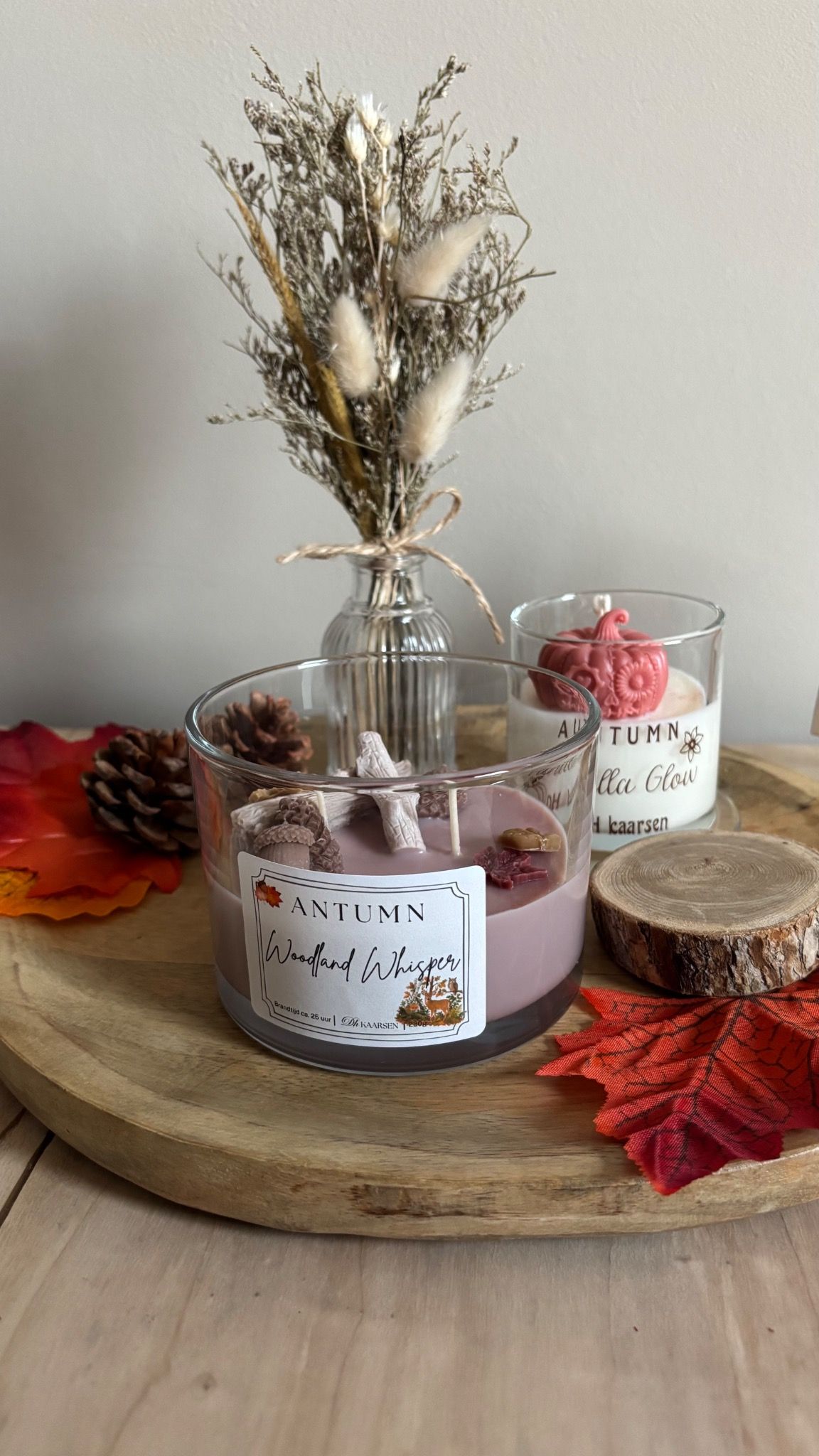 Glazen kaars met gedroogde bloemen op een houten schaal, versierd met herfstbladeren op een lichte houten tafel.