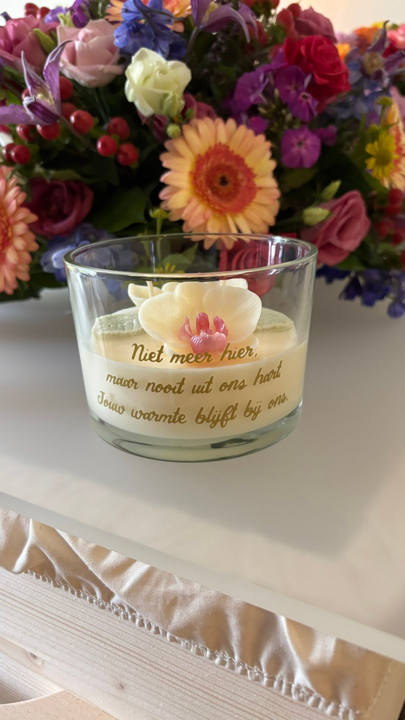 Een kaars in een doorzichtige glazen pot met een sierlijk etiket, geplaatst voor een kleurrijk boeket op een witte tafel. rouwkaars