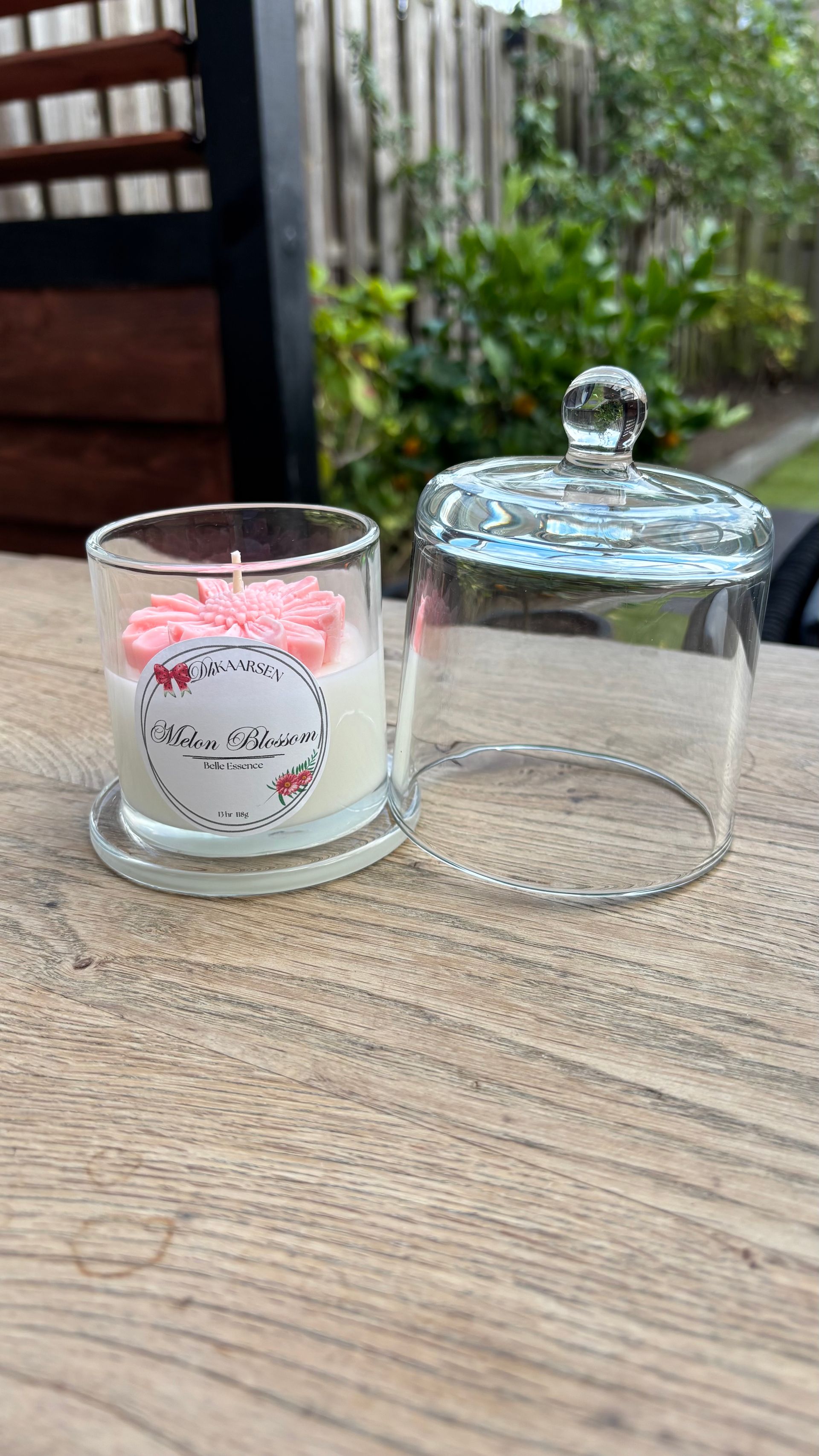 Glazen kaarsenhouder met roze bloemen naast een doorzichtig glazen deksel op een gevlochten tafel buiten.