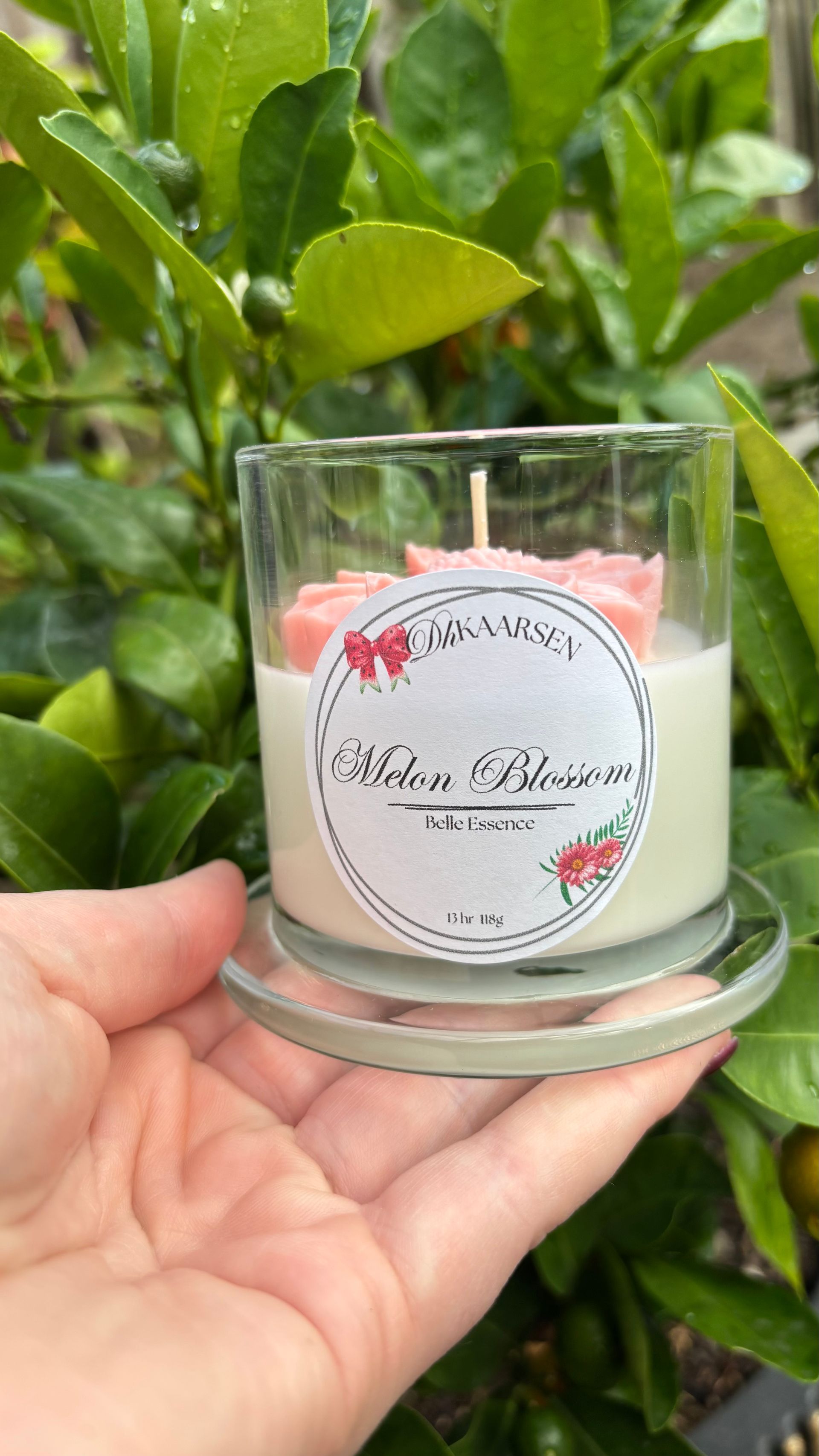 Glazen kaarsenhouder met roze bloemen naast een doorzichtig glazen deksel op een gevlochten tafel buiten.