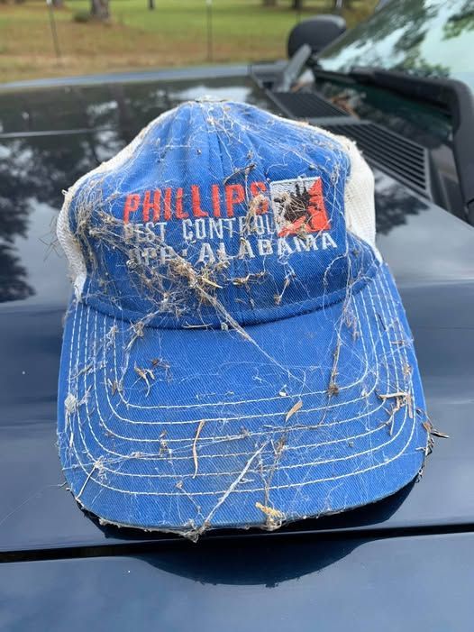 Blue worn Phillips “East Central Alabama” baseball cap on a car hood, covered with grass and debris