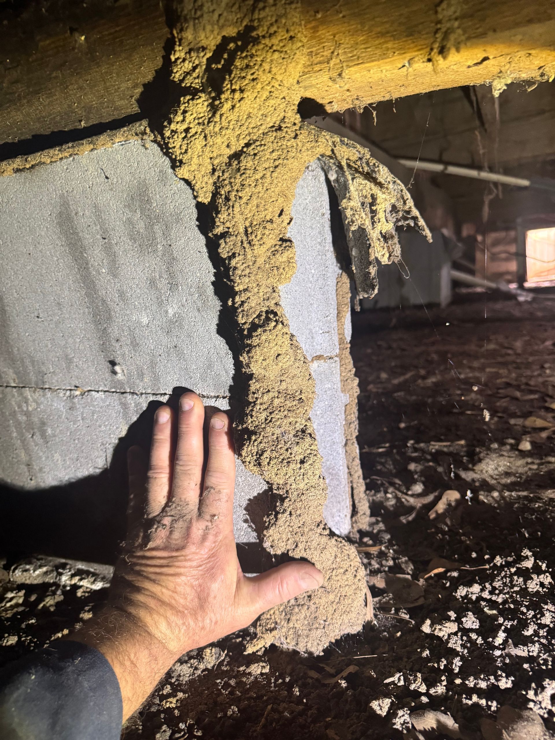 Hand touching a damaged concrete pillar with exposed yellow foam and debris in a dark crawlspace