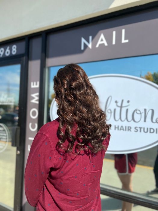 Person with long curly hair in a red sweater facing a nail salon storefront window.