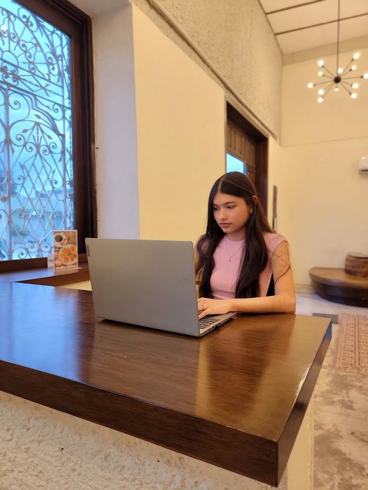 A person sitting at a wooden counter in a bright room, typing on a laptop, with a large window and chandelier nearby.