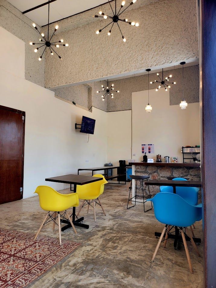 Interior with cafe tables and colorful chairs, under globe-shaped lights. Concrete floor, high ceilings.