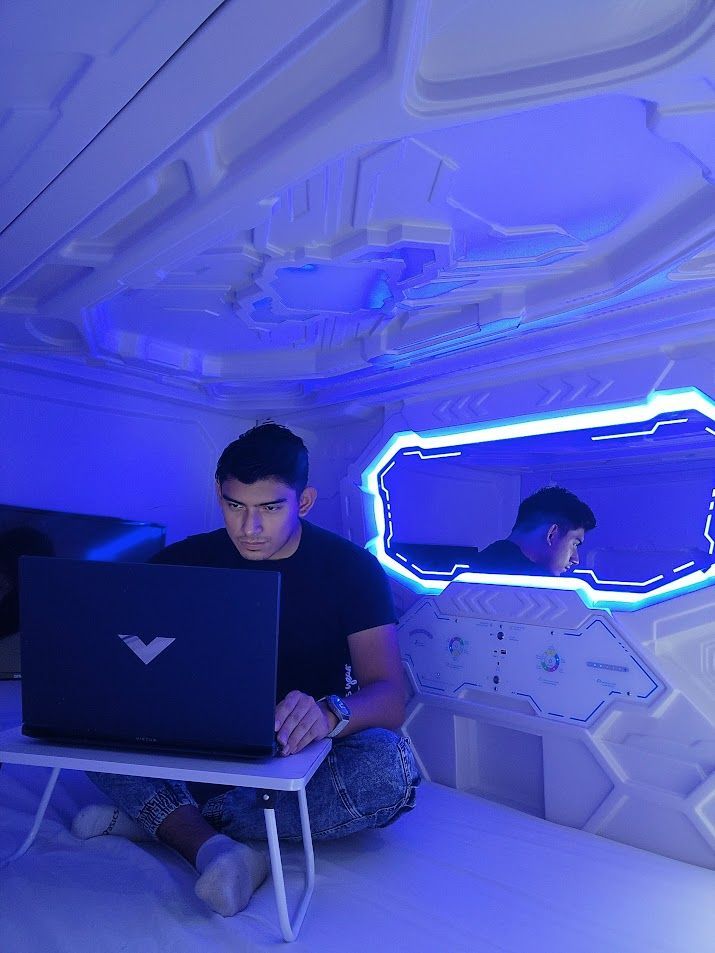 Man working on a laptop in a futuristic capsule lit by blue light.