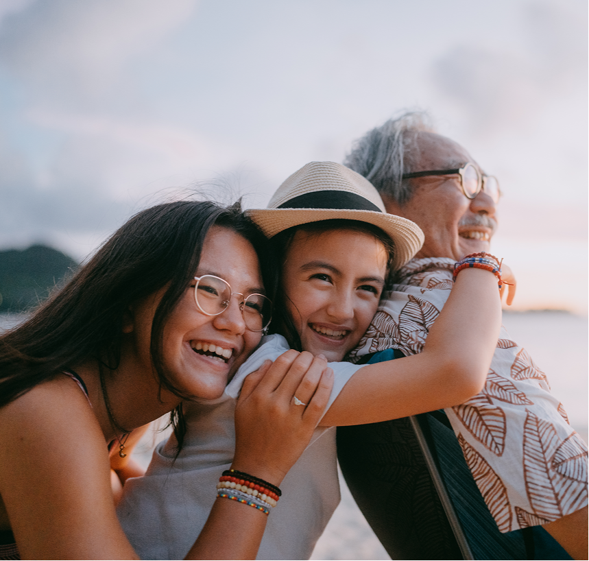 Three smiling friends outdoors at sunset, leaning together and laughing on a beach.
