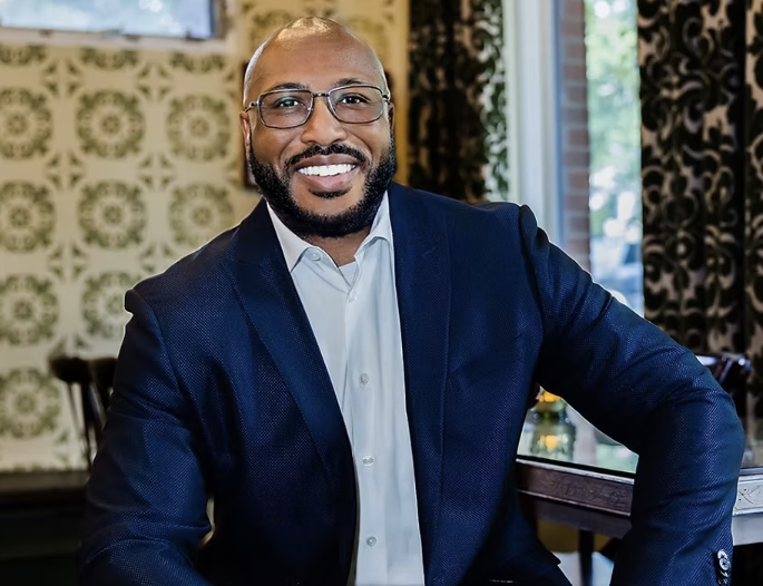 Smiling man in navy blazer seated indoors with patterned wallpaper and a window behind him