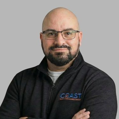 Portrait of a bald man with glasses and a black zip-up jacket against a gray background