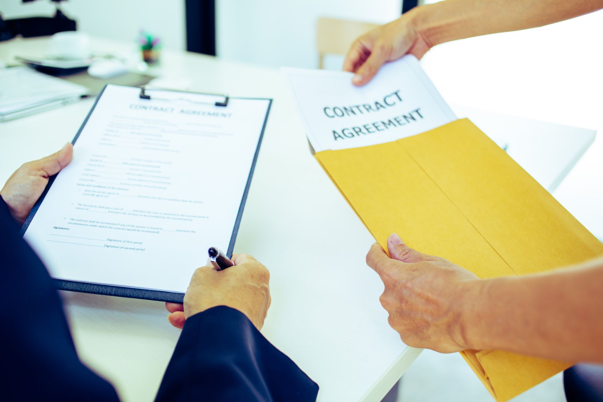Hands exchanging contract documents, one person holding a clipboard and pen, indoors at a desk.