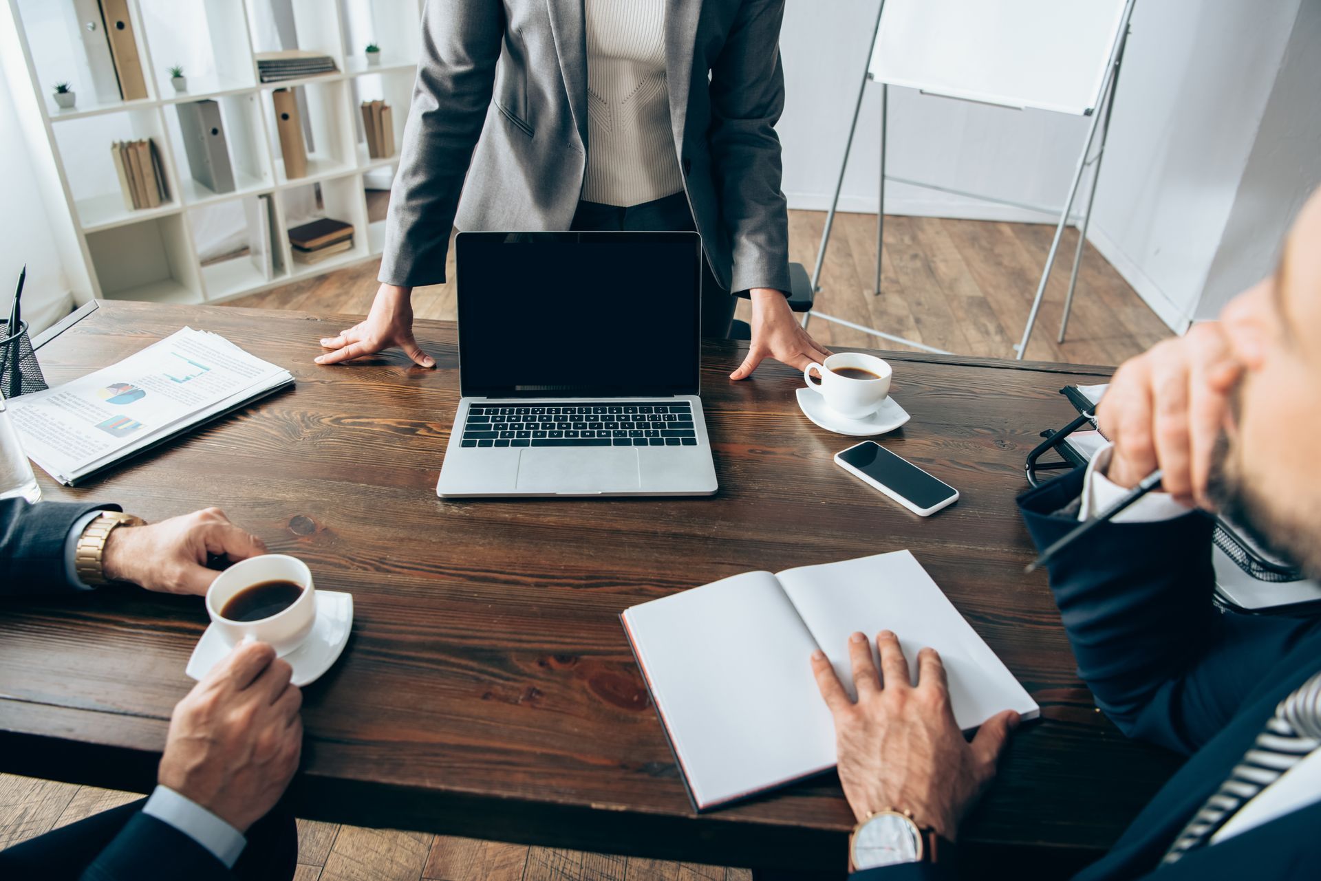 Business meeting: three people around a table, one standing with hands on laptop, others seated with coffee and notepad.