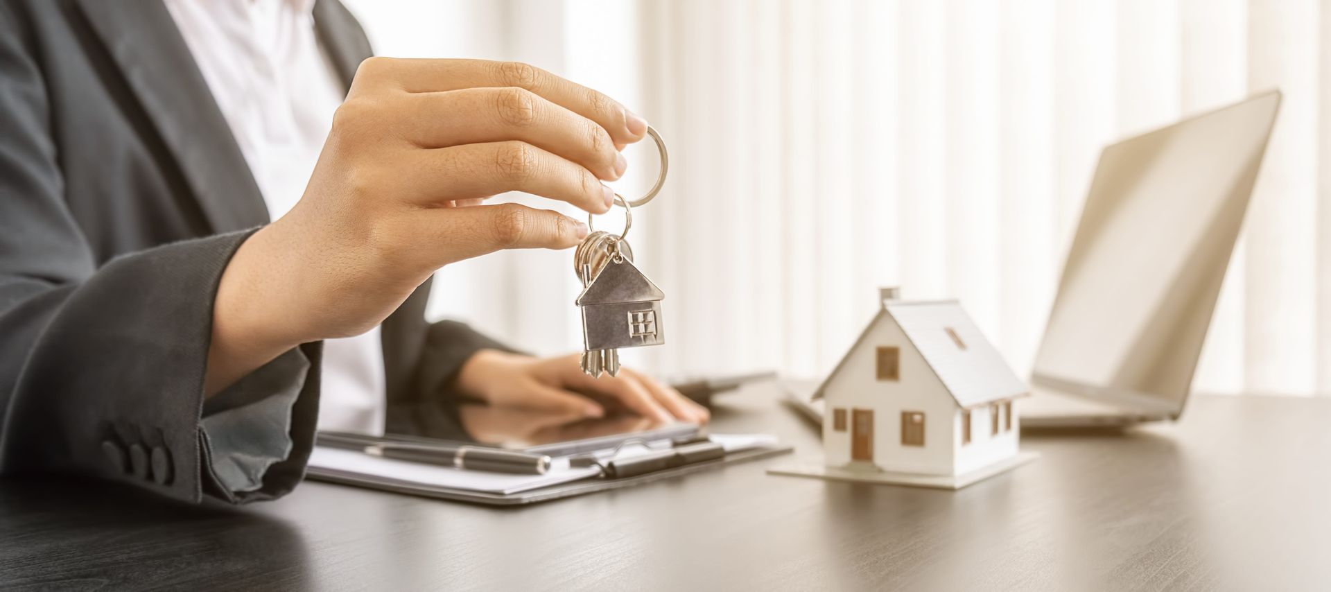 Hand placing a key beside a small house model and tablet on a desk, with a laptop in the background