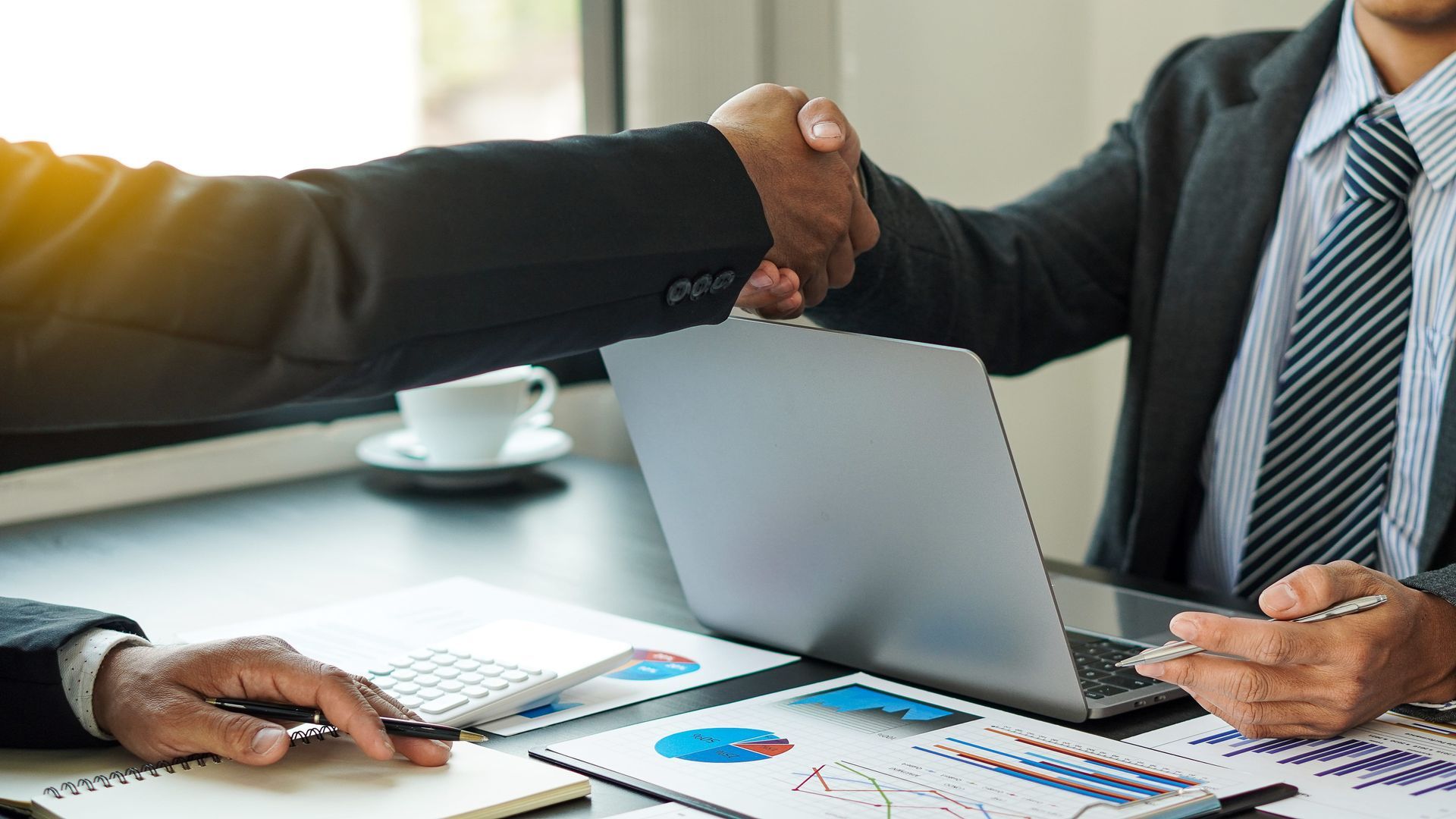 Two businesspeople shake hands across a laptop at a meeting table with charts and coffee cups.