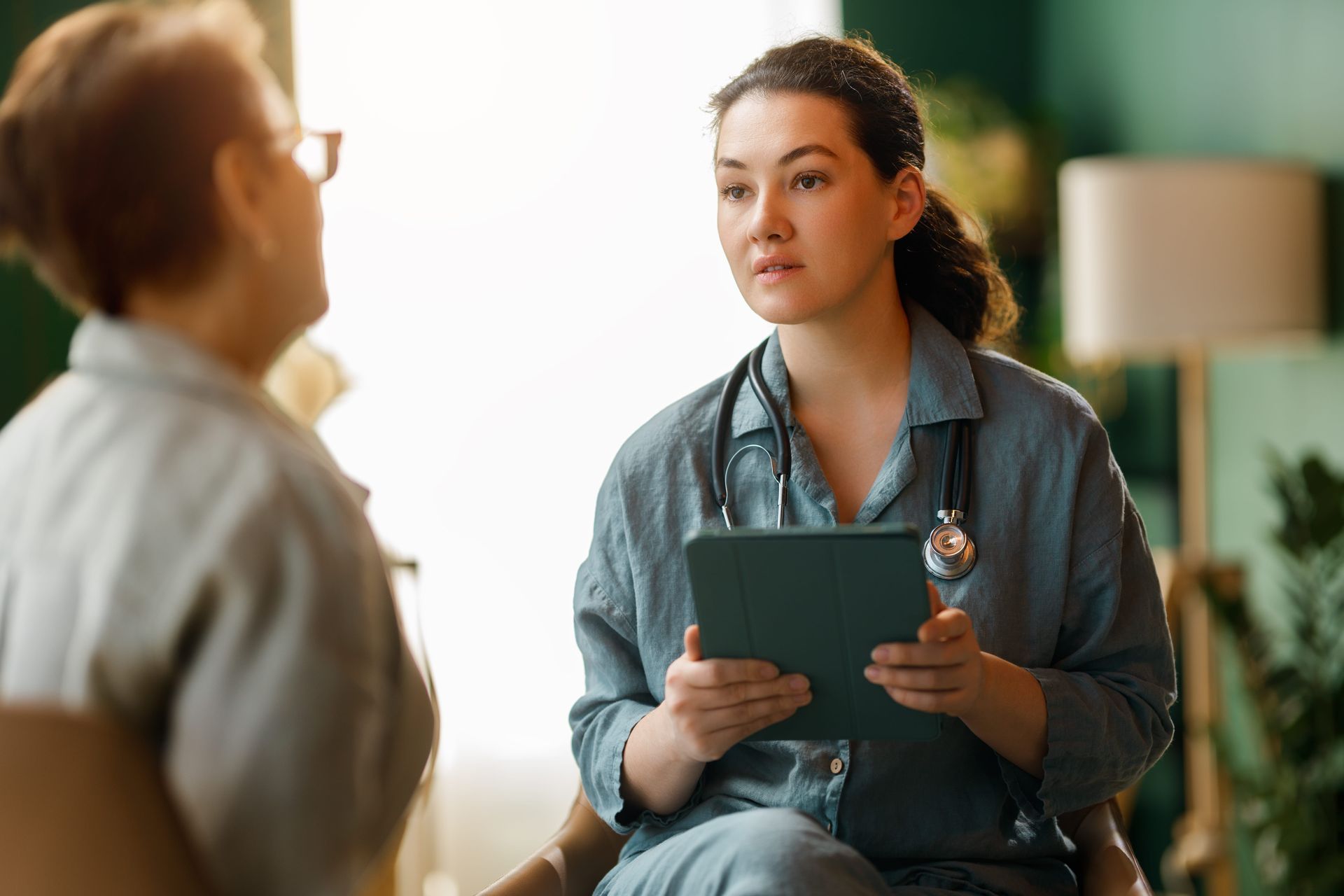 Healthcare worker holding a tablet while speaking with a seated patient in a clinic.