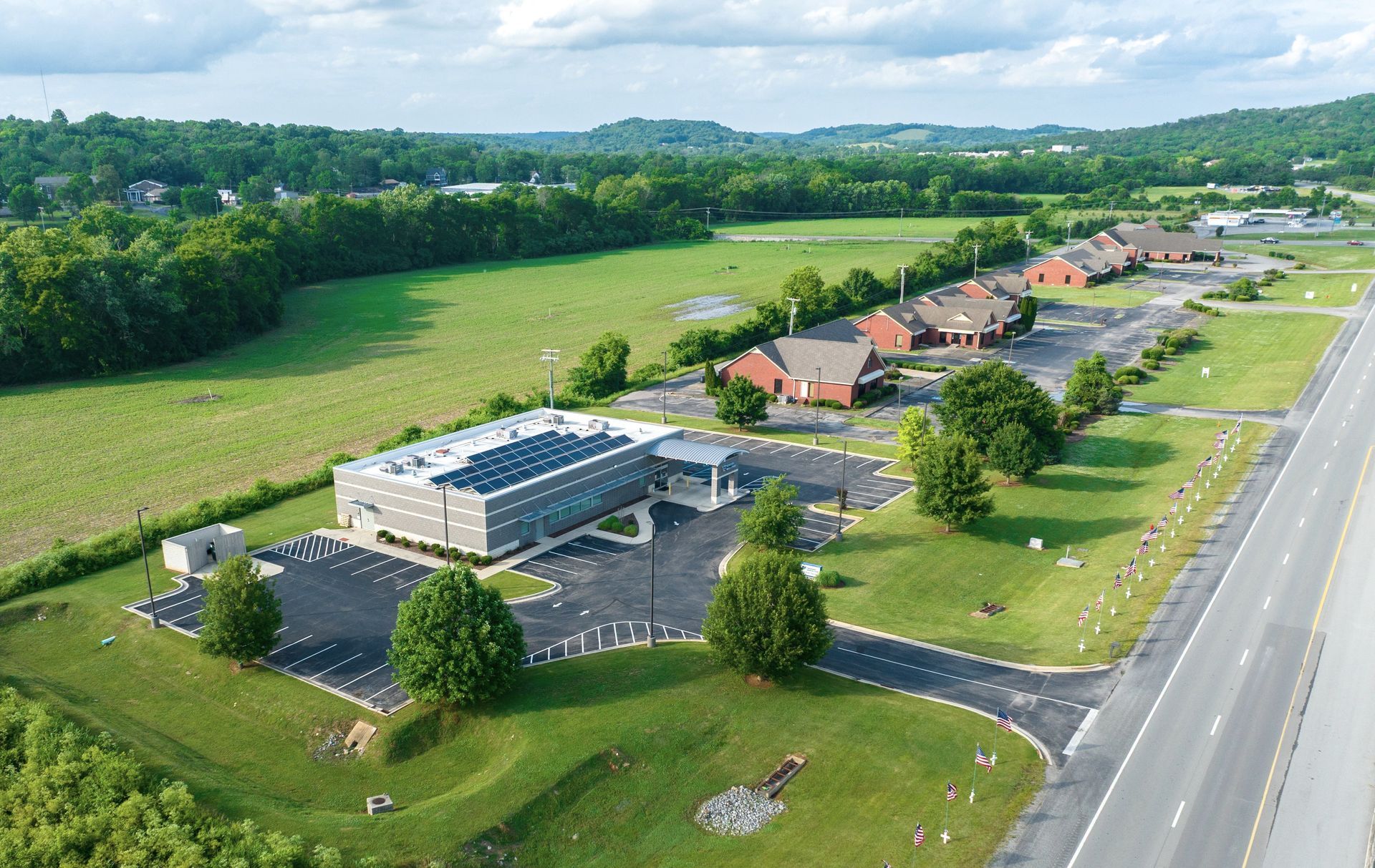 Aerial view of a Fresenius Medical Care building complex beside a highway, surrounded by green fields and trees.
