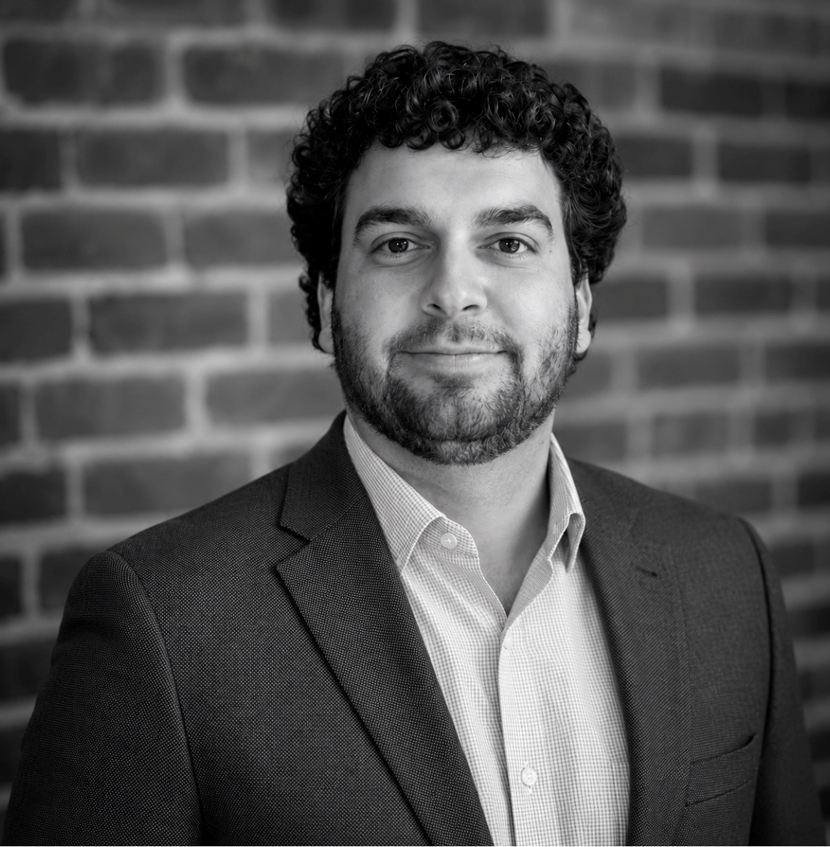 Black-and-white headshot of a Joseph Brown in a blazer smiling slightly against a brick wall.