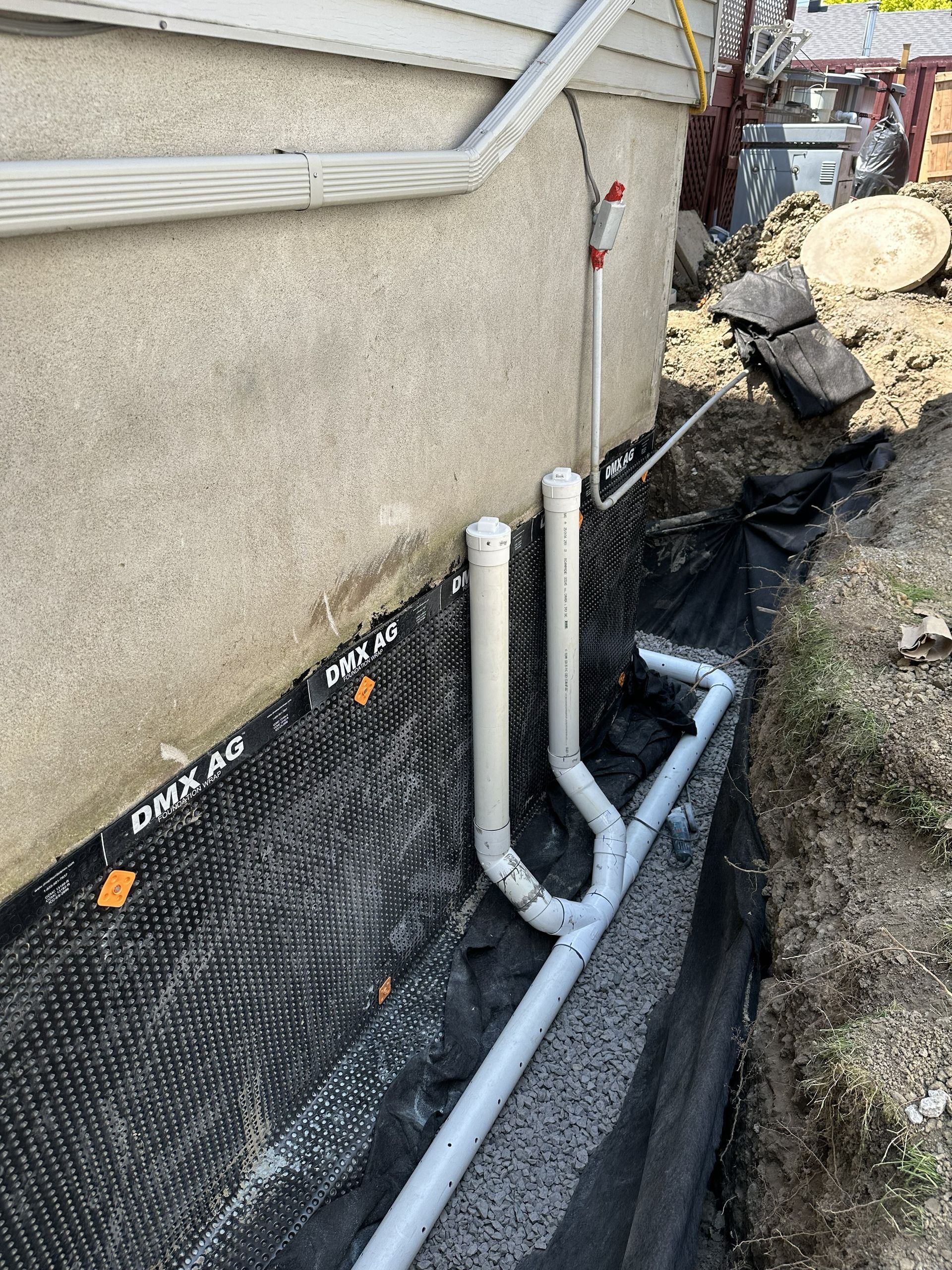 Une tranchée le long d'un mur de fondation, avec une membrane noire à picots et des tuyaux en PVC blanc reliés à un système de drainage.