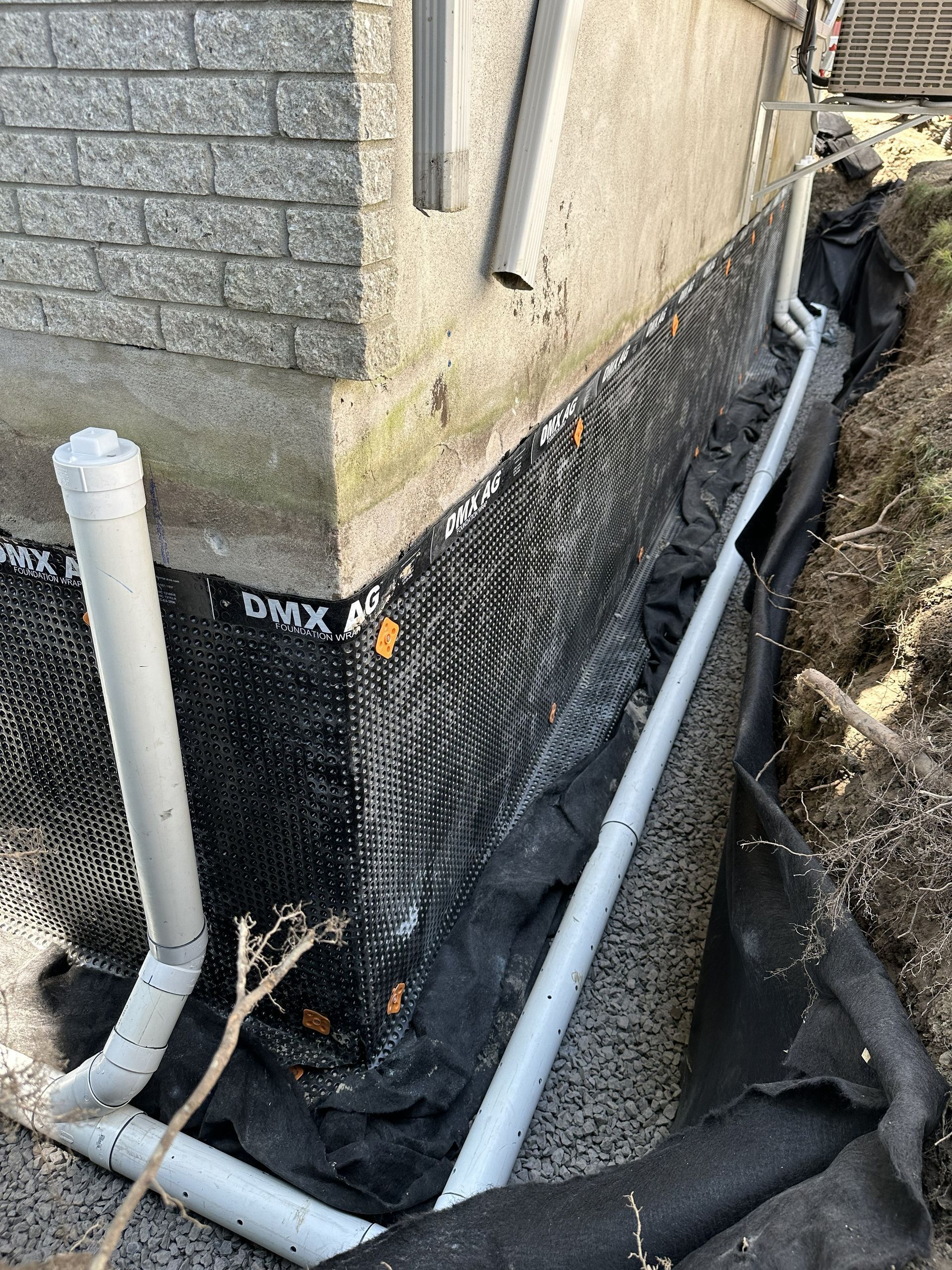 Un mur de fondation a été excavé et équipé d'une membrane de drainage noire à picots et d'un nouveau tuyau de drainage périphérique en PVC gris.