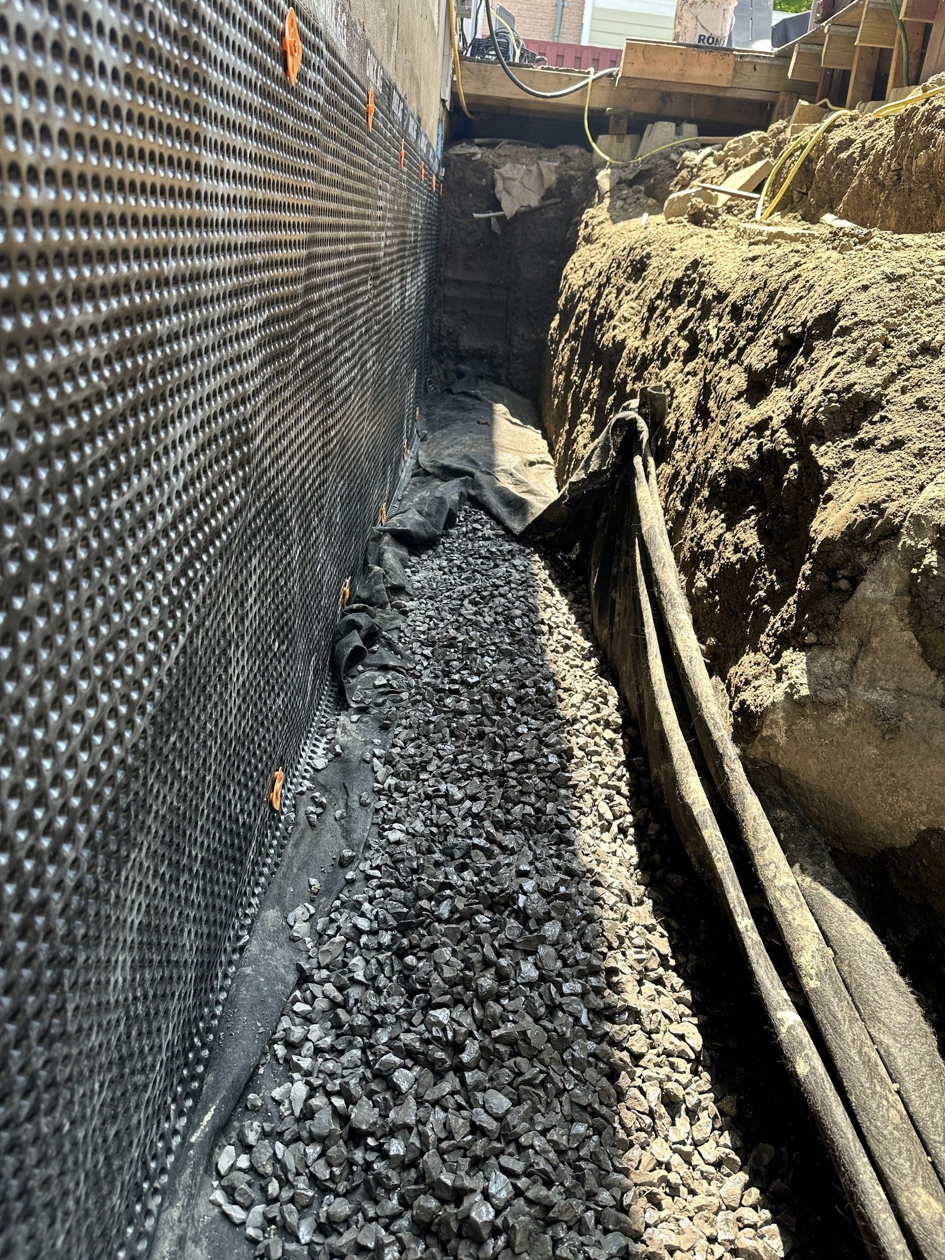 Un mur de fondation recouvert d'une natte de drainage noire jouxte une tranchée remplie de gravier et de tuyaux de drainage.