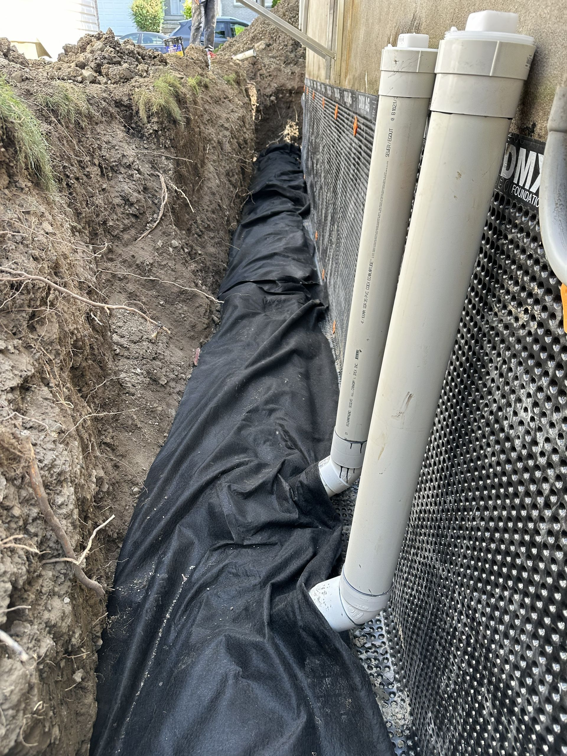 Une tranchée le long des fondations d'un bâtiment, avec un géotextile noir recouvrant les tuyaux de drainage et une dalle de fondation alvéolée.