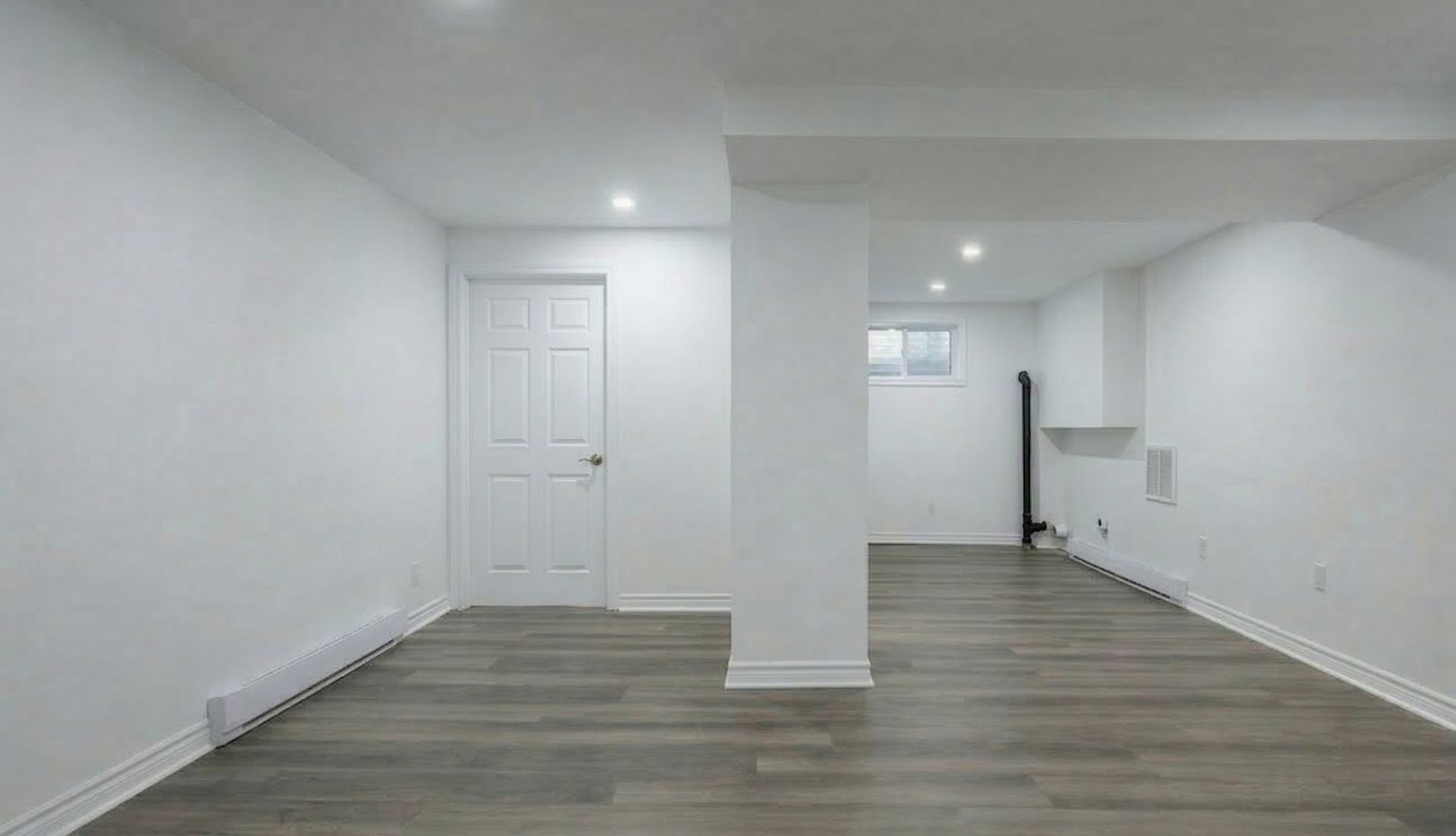 Une pièce minimaliste et vide au sous-sol, avec des murs blancs, un sol gris imitation bois, une simple porte blanche et un éclairage encastré.