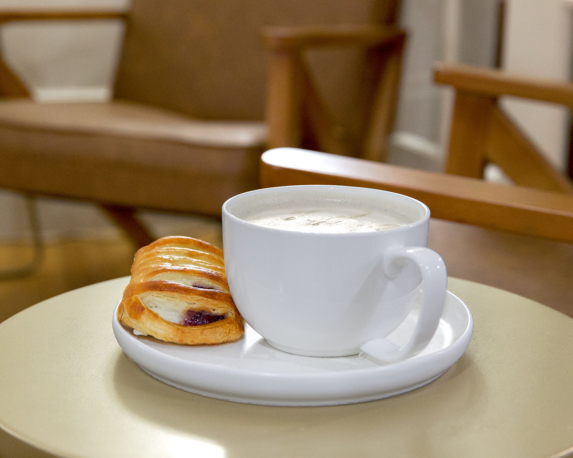 White coffee cup with latte on saucer beside a small pastry on a table in a cozy café